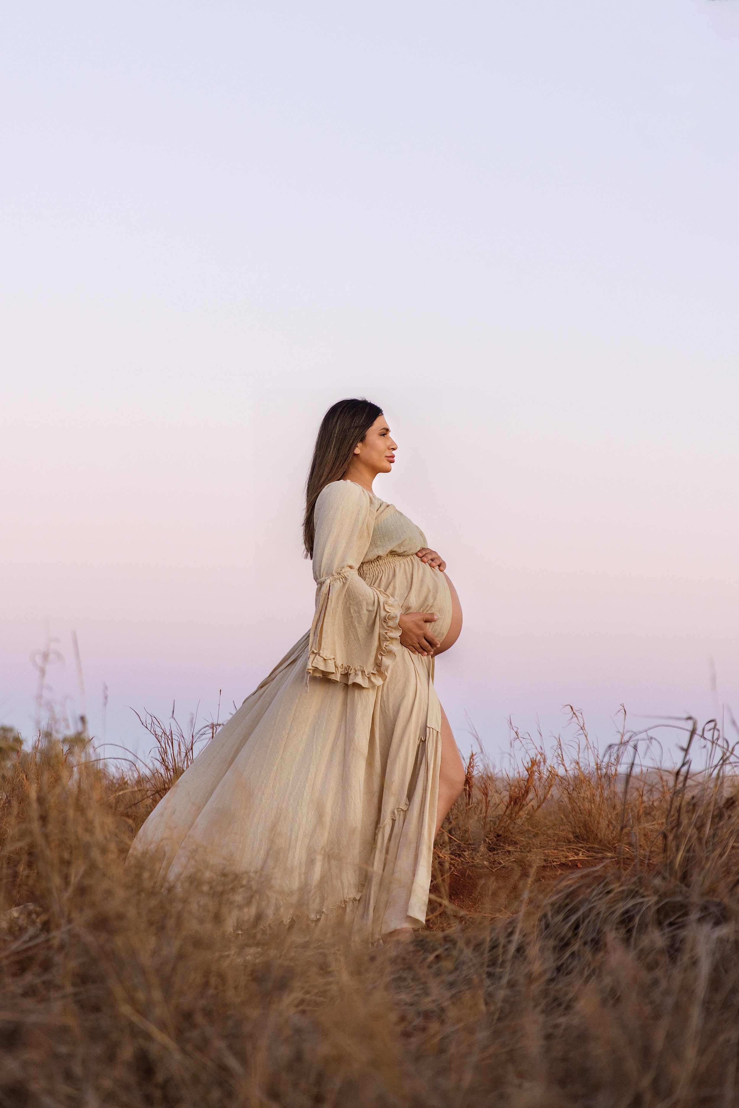 Maternity session, photography, Photographer, photoshoot. Pregnant woman in a flowing beige dress walking through tall dry grass outdoors during sunset or sunrise