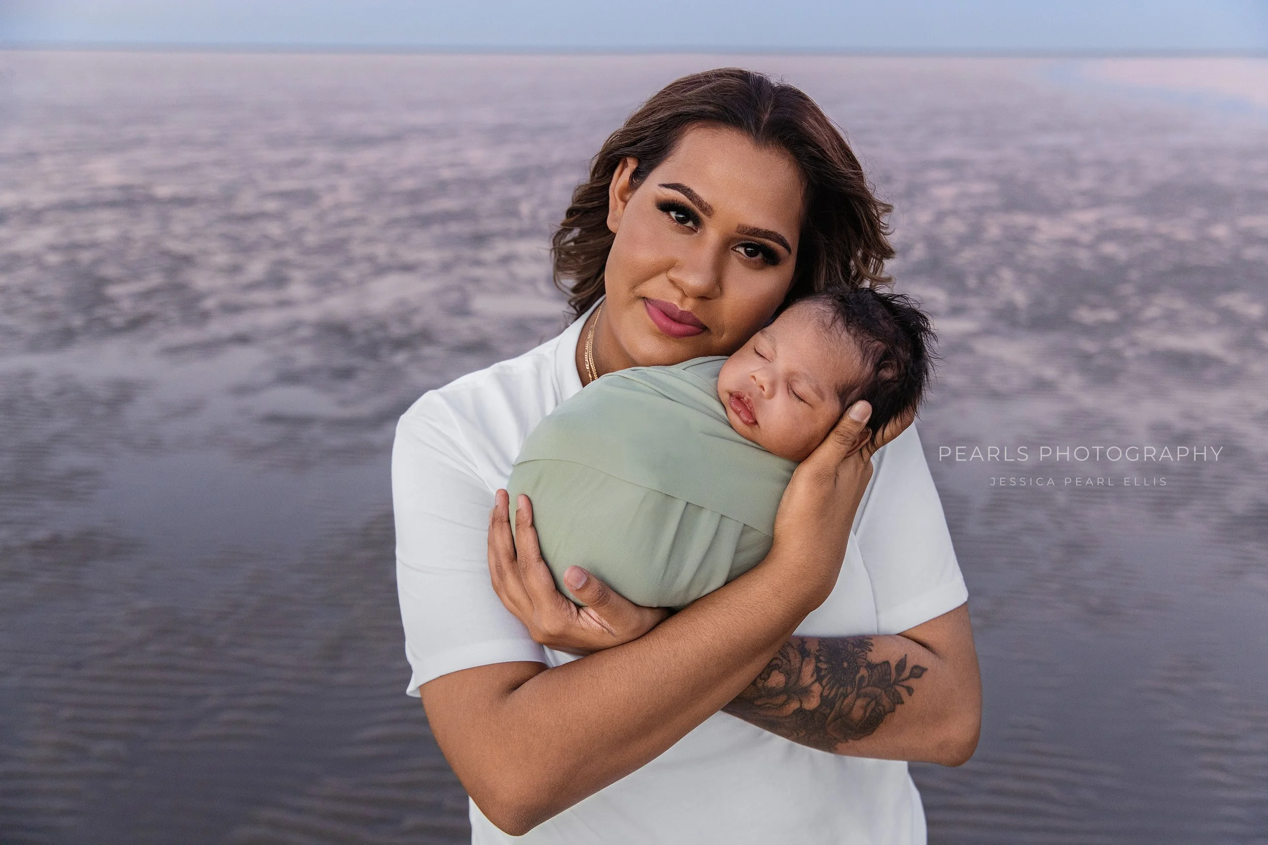 A woman with medium skin tone and tattoos on her arm holding a sleeping child with dark hair by a body of water, possibly a lake or ocean, during sunset or dusk. Outdoor newborn photoshoot, Karratha photographer
