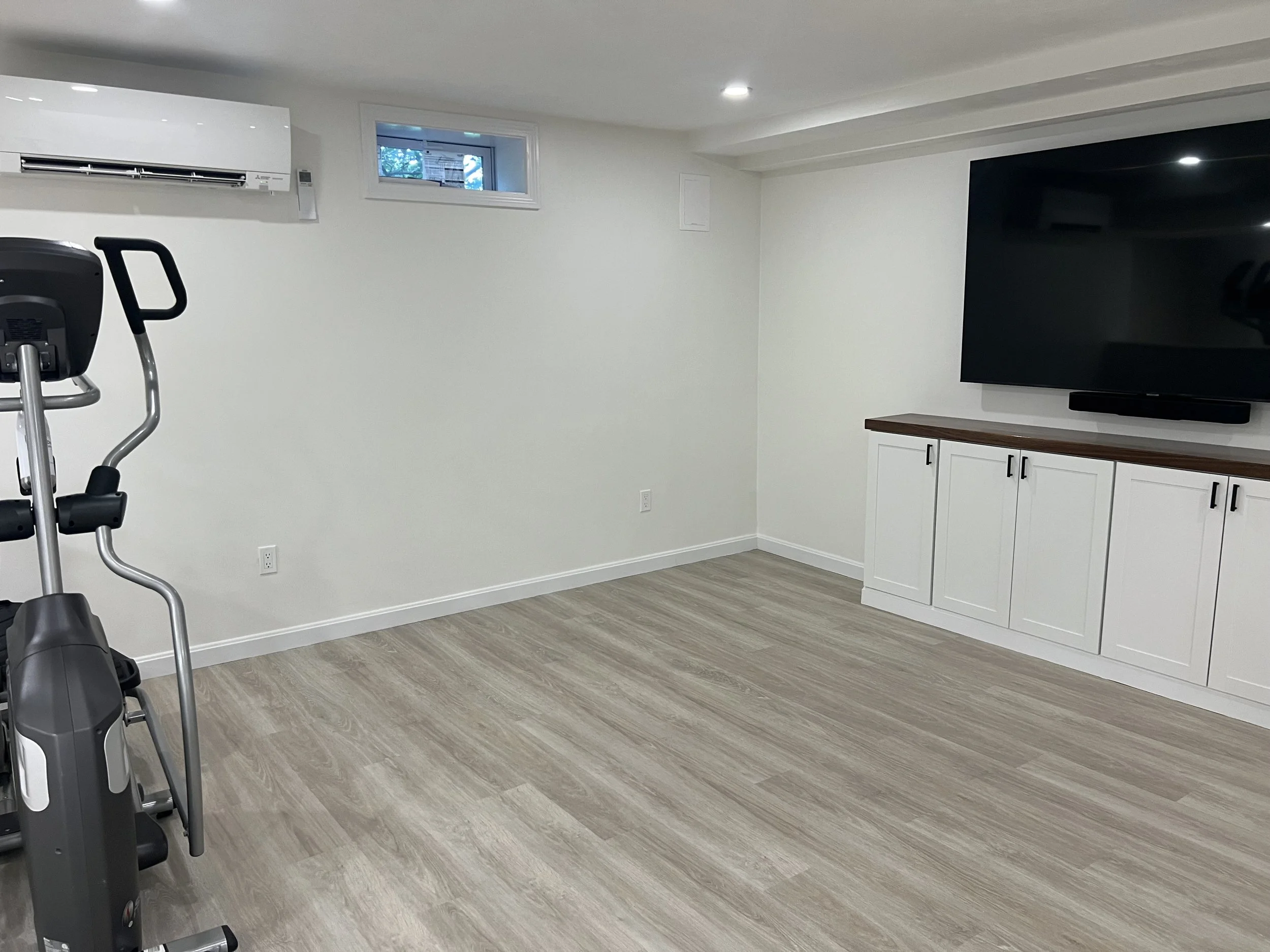 Empty basement room with gym equipment, a large TV, white cabinets, and a small window in the wall.