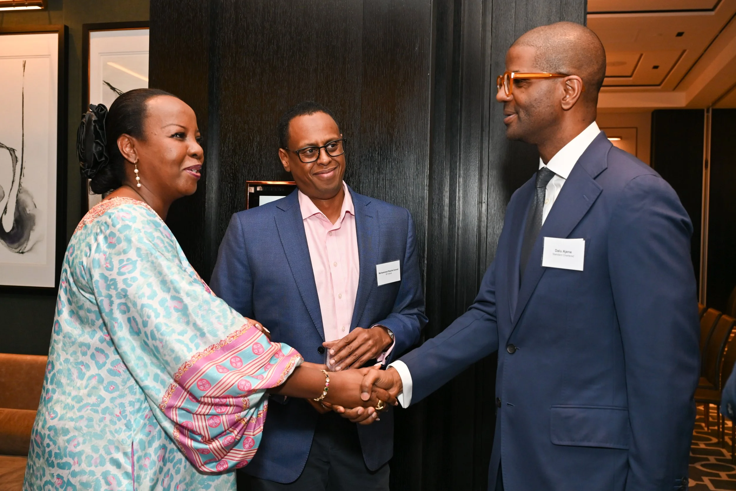 Three people are engaged in a handshake at a formal event. The woman on the left is wearing a patterned dress and jewelry, the man in the middle is dressed in a blue suit with glasses, and the man on the right is wearing a dark suit, tie, and glasses. They are standing in a room with black walls and framed artwork.