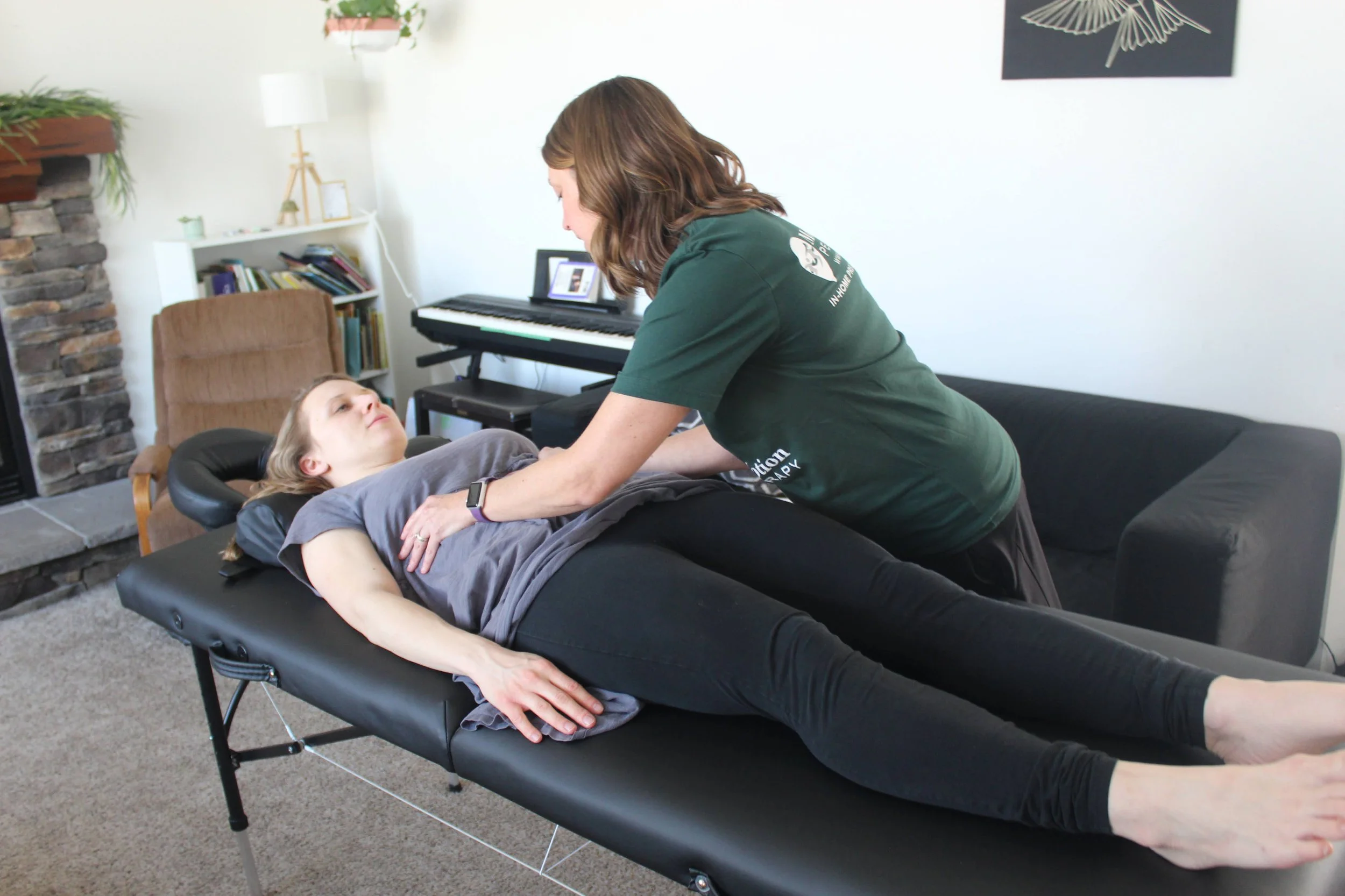 A woman providing physical therapy to a young woman lying on a portable therapy table in a living room.