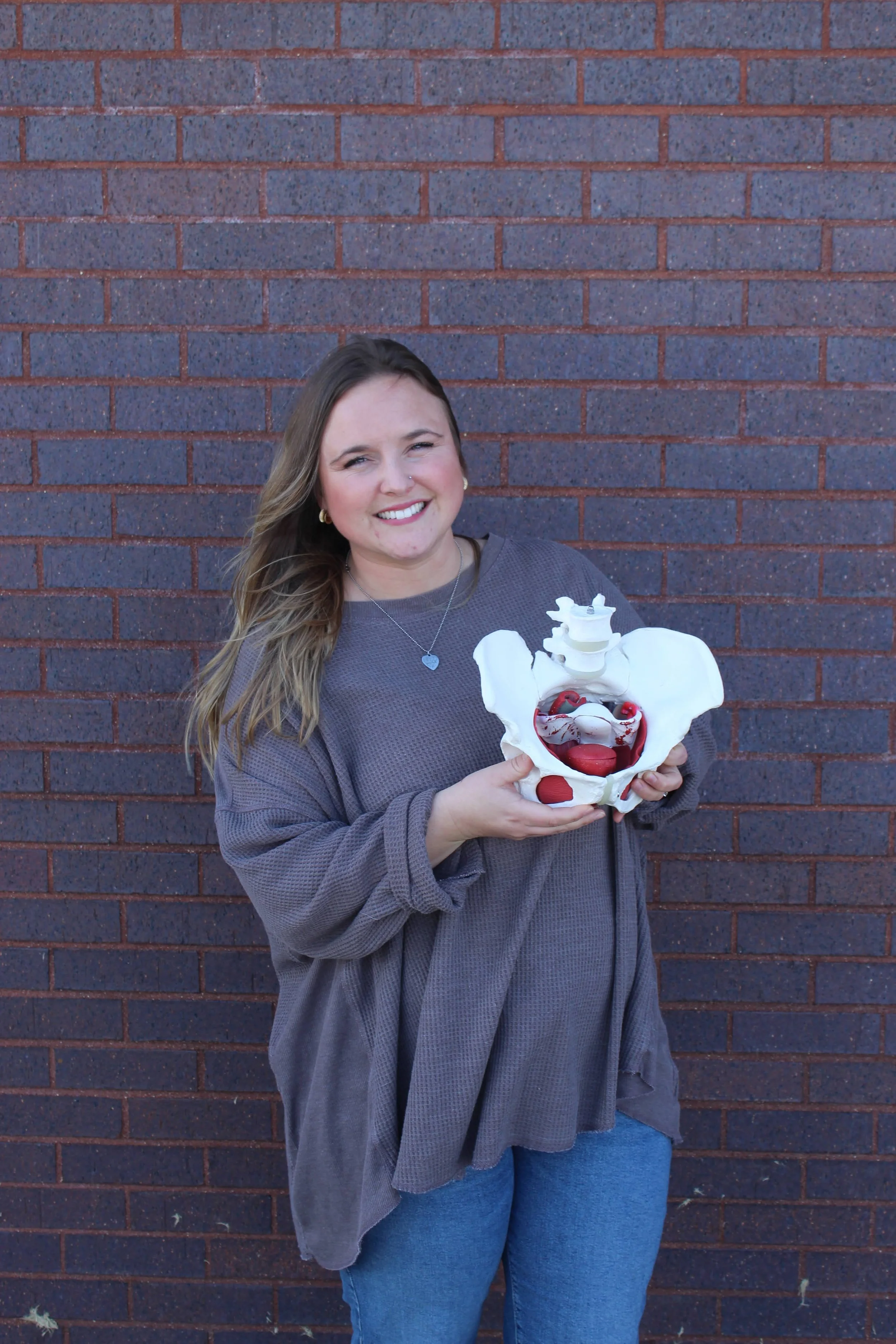 A woman with long hair smiling while holding a model of a human pelvis and reproductive organs against a brick wall background.