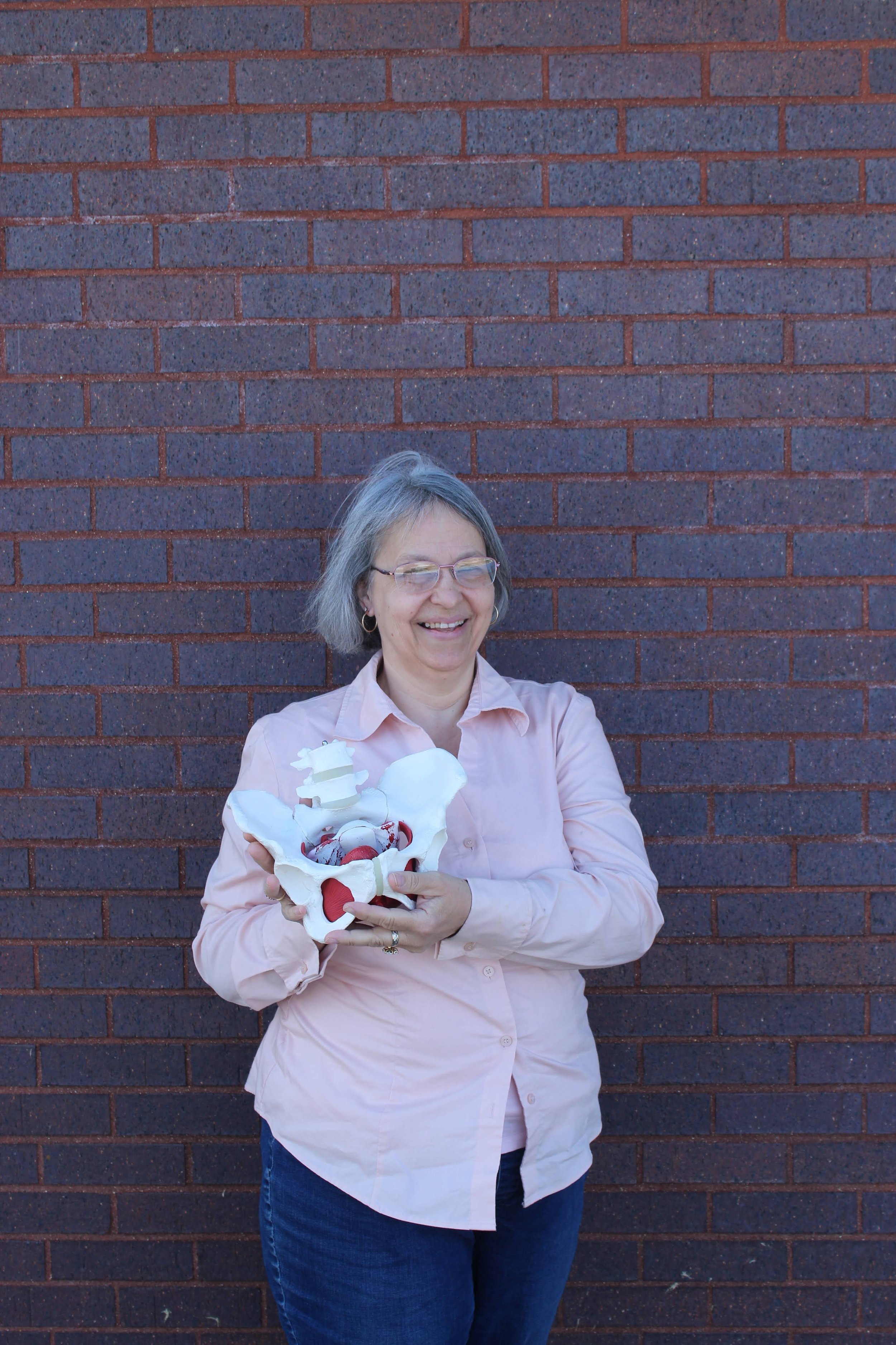 A woman with short gray hair and glasses smiling while holding a model of a pelvis with spinal and heart decorations, standing against a brick wall.