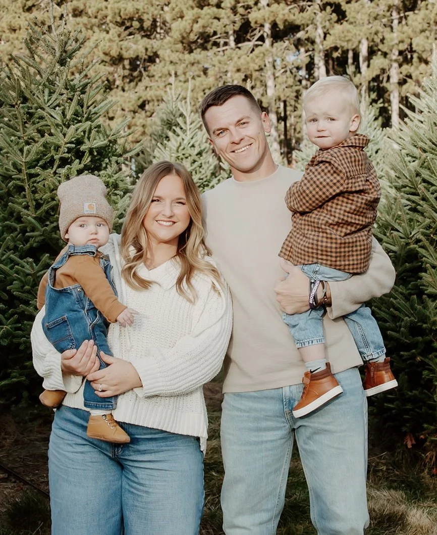 A family of four standing outdoors among Christmas trees, smiling at the camera. The woman has long blond hair and is holding a young boy with blond hair, dressed in a brown jacket and jeans. The man has short dark hair and is holding a young girl in a brown and black checkered shirt and jeans. The background features numerous green Christmas trees and a forested area.