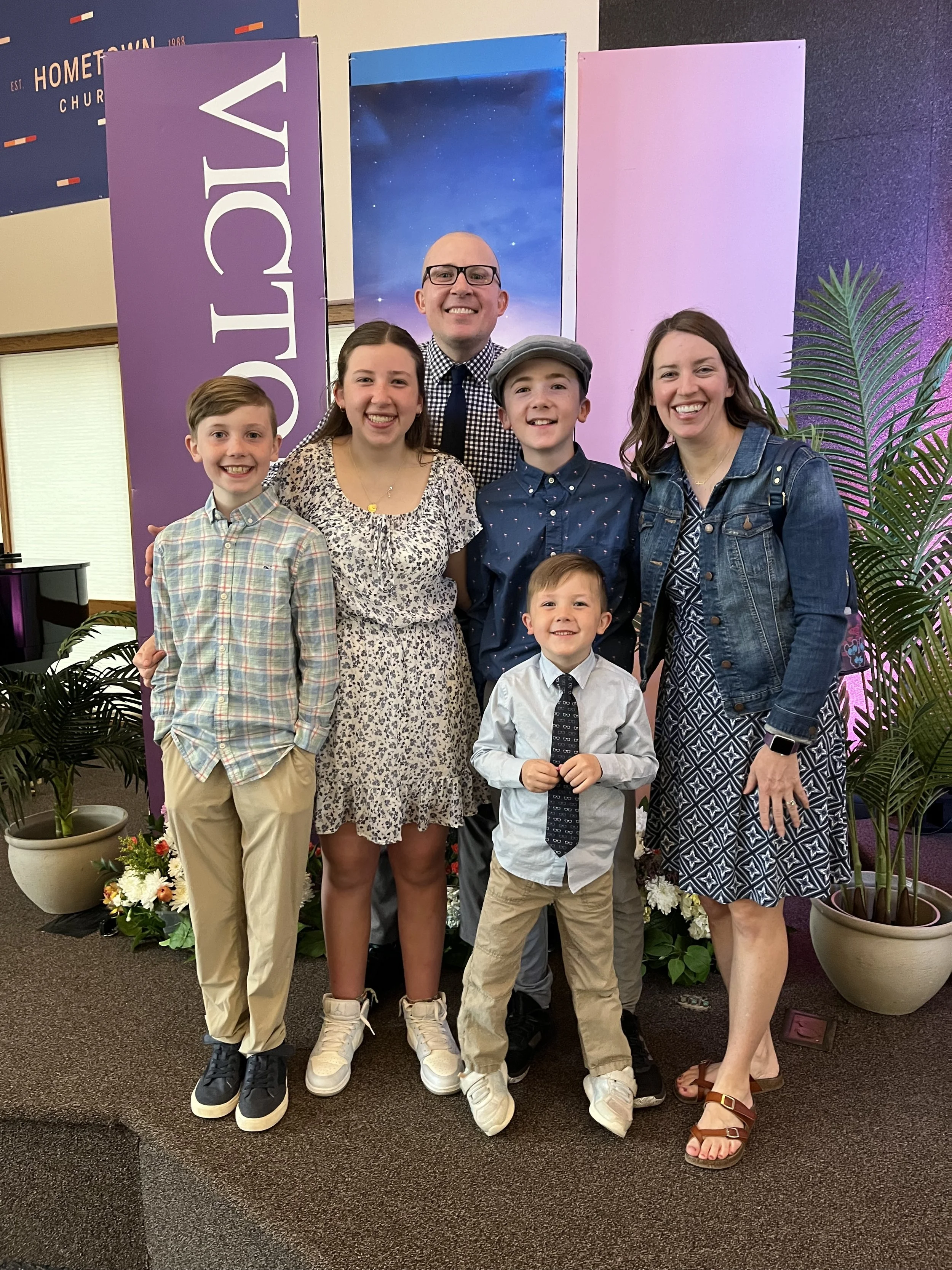 A group of seven people, including two adults and five children, smiling and posing together in a church setting with purple and pink panels, plants, and flowers in the background.