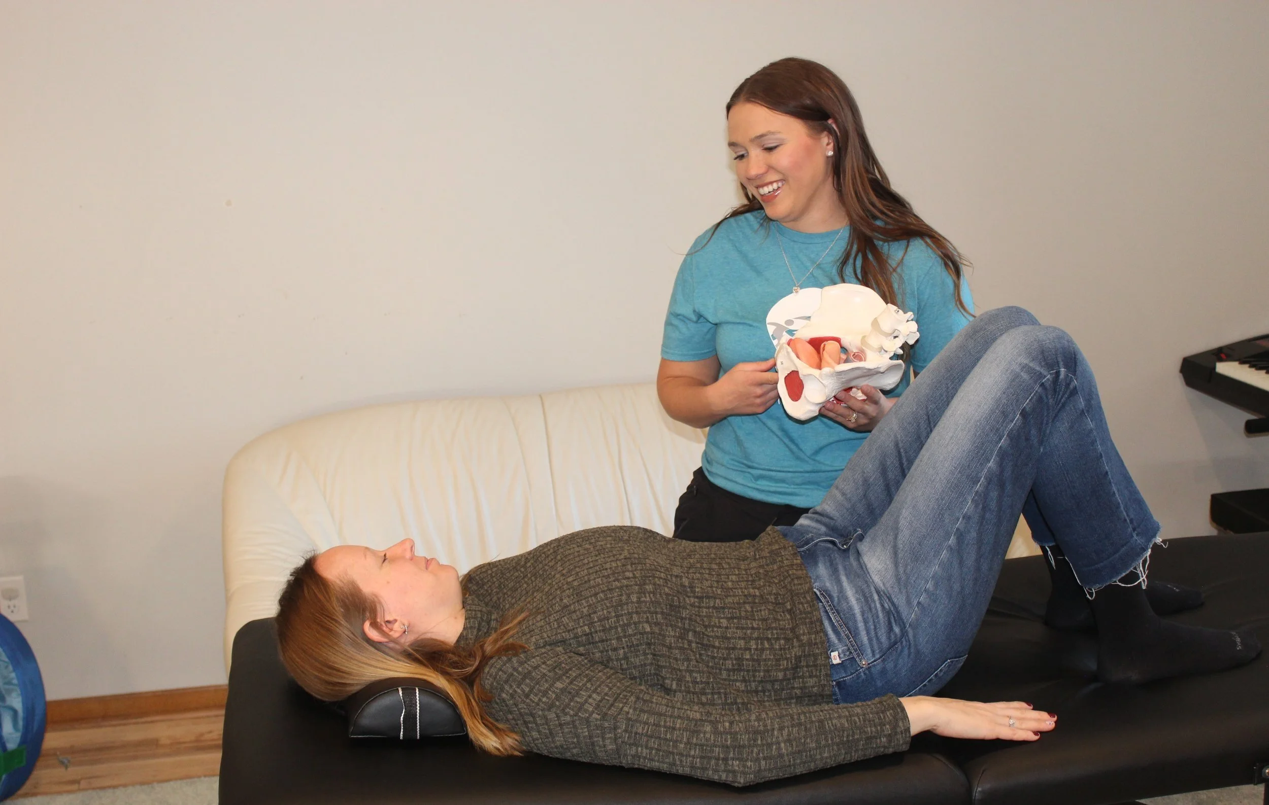 Two women practicing pelvic physical therapy, one lying on a treatment table with legs raised and the other standing, holding a model of the pelvic region.