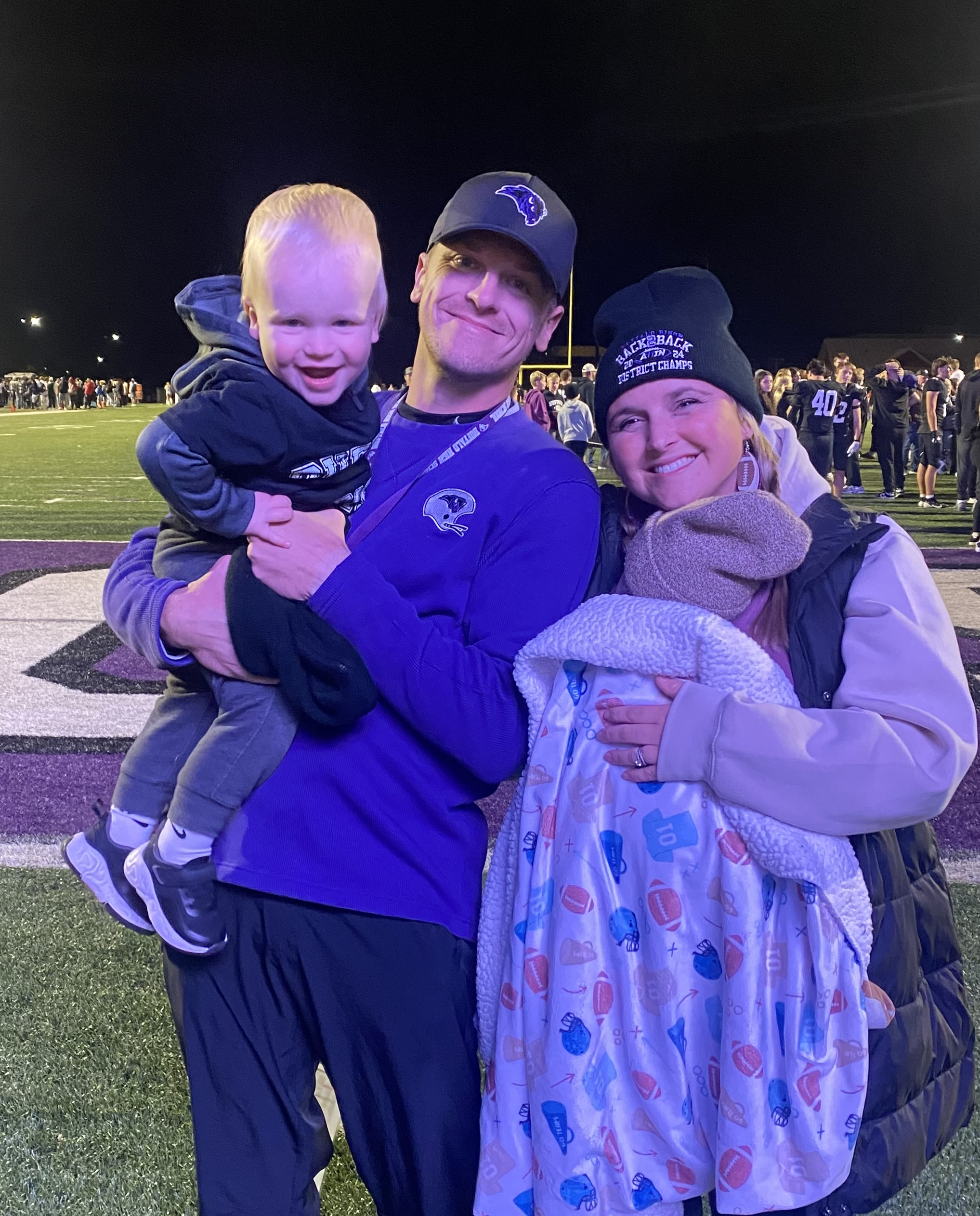 A group of three people on a football field at night, smiling and embracing. A man in a dark jacket holds a young blond boy, and a woman in a beanie and coat stands beside them with a blanket.