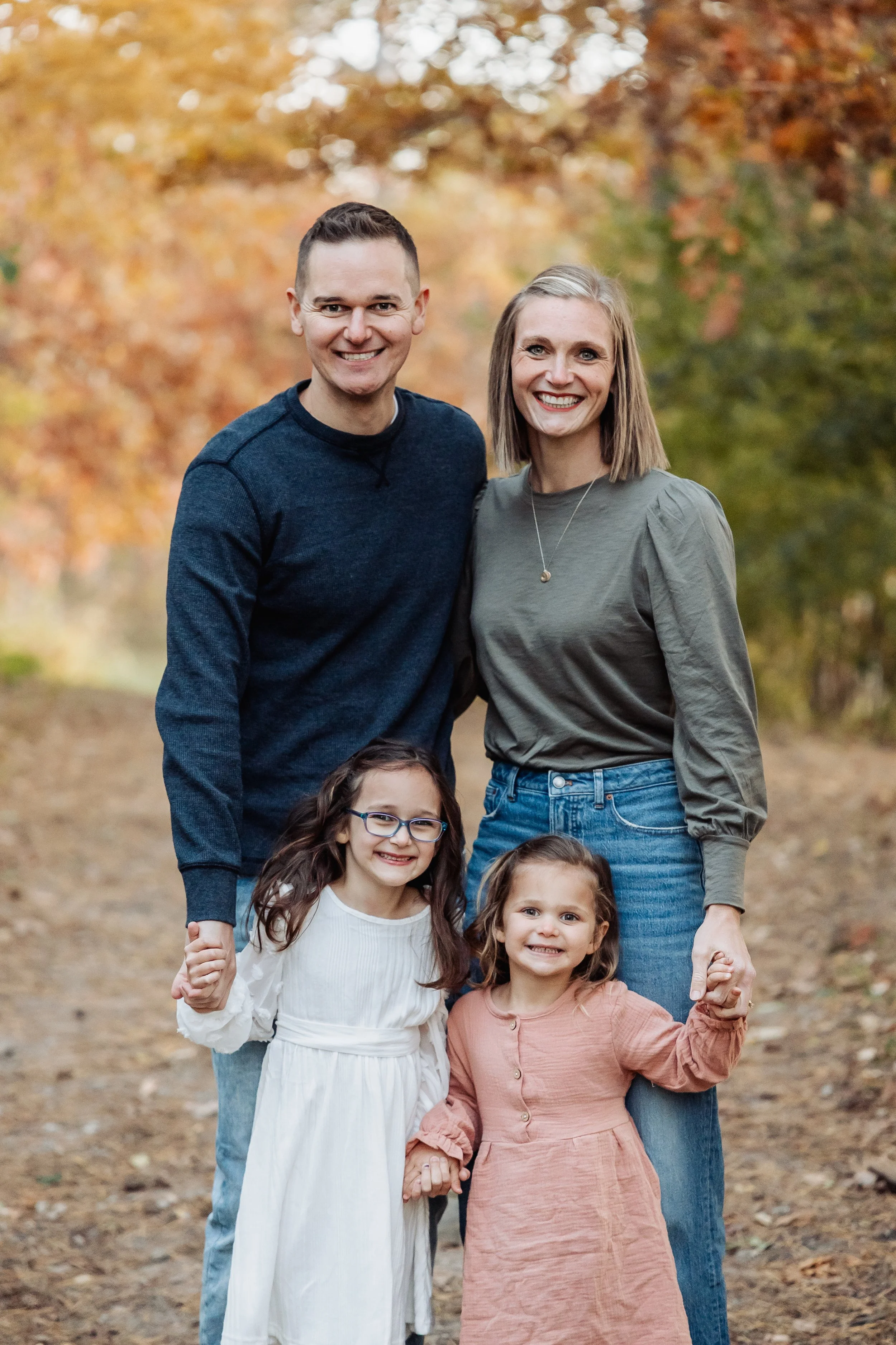 A happy family of four holding hands outdoors in fall, with autumn trees in the background.