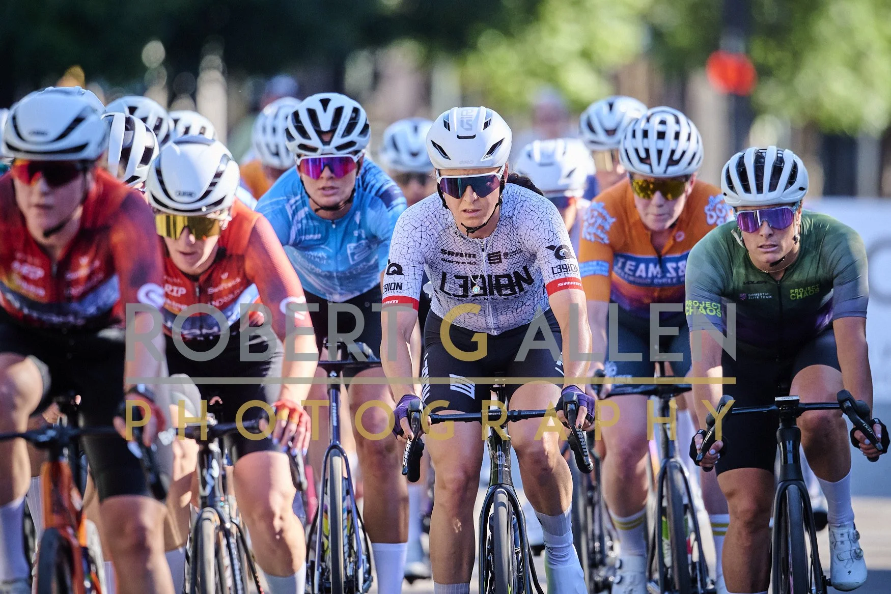 A group of female cyclists racing on a city street, wearing helmets and colorful jerseys.