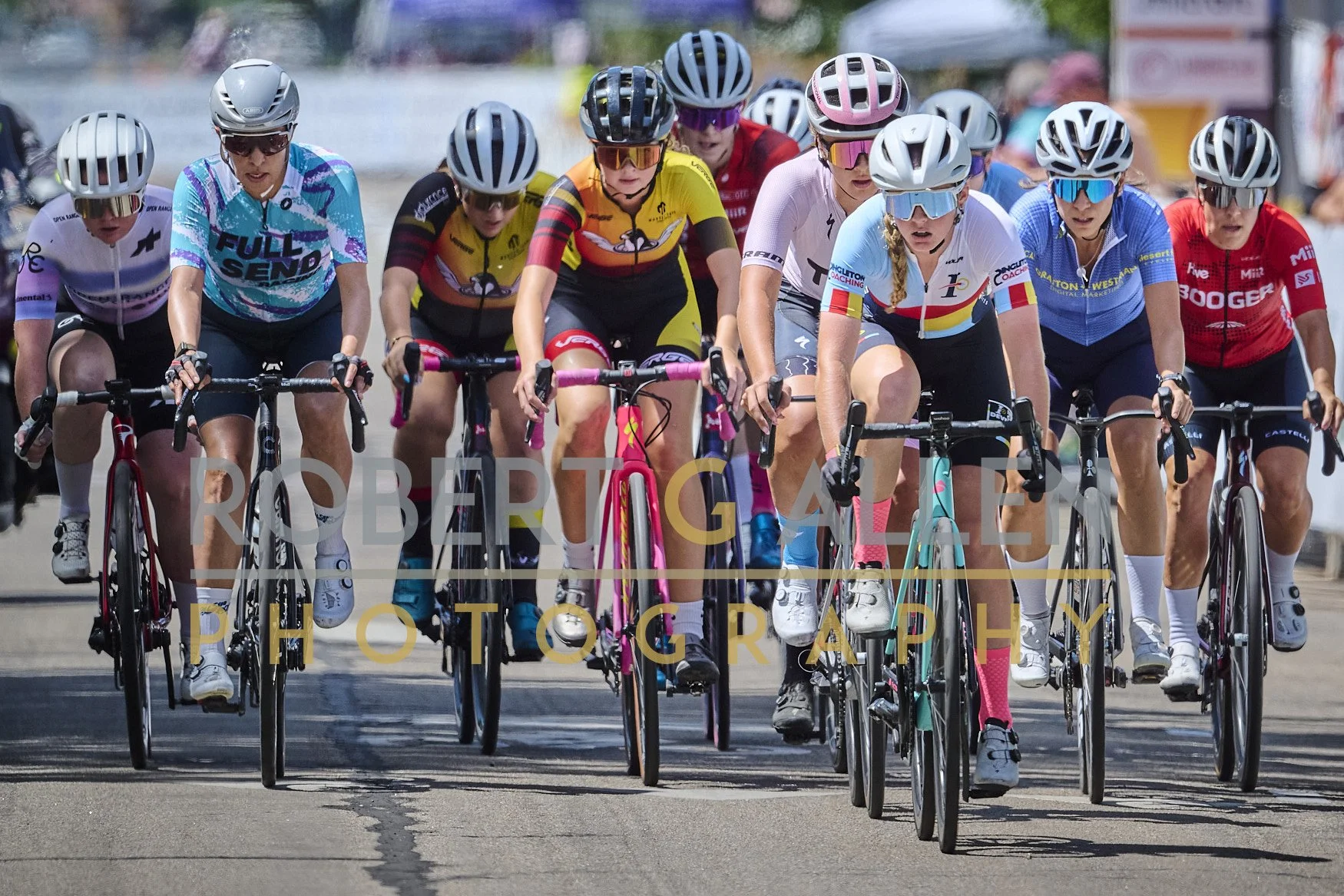 A group of female cyclists participating in a race, riding closely together on a paved road during the daytime, with some wearing colorful jerseys, helmets, and sunglasses.