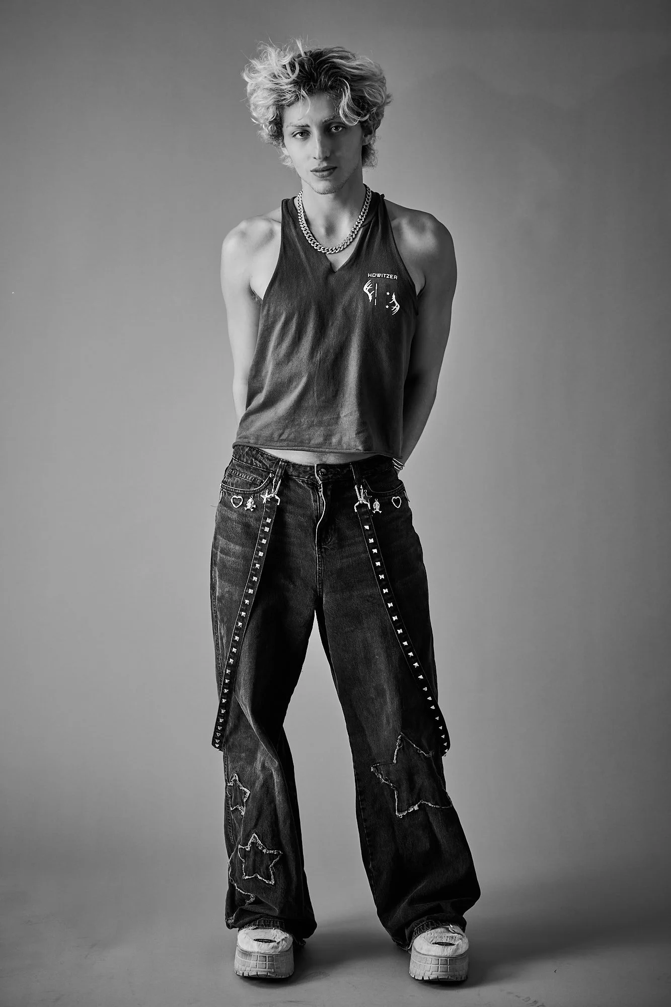 Black and white photo of a young person with curly hair, wearing a sleeveless top, baggy jeans with star patches and rips, chunky sneakers, and chains around their neck. They are standing against a plain background, facing forward, with hands behind 