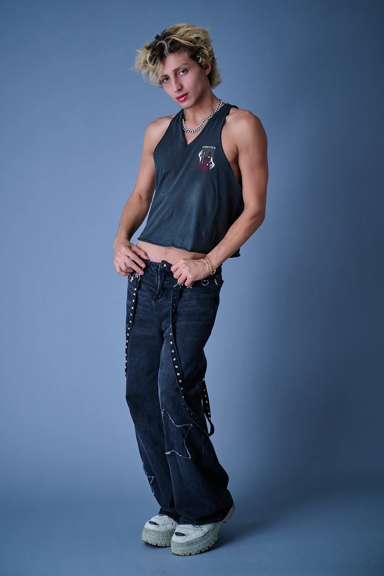 A young person with blonde curly hair posing against a plain blue background, wearing a black sleeveless top, baggy black jeans with chain details, chunky white sneakers, a gold bracelet, and a chunky silver necklace, with a confident expression.