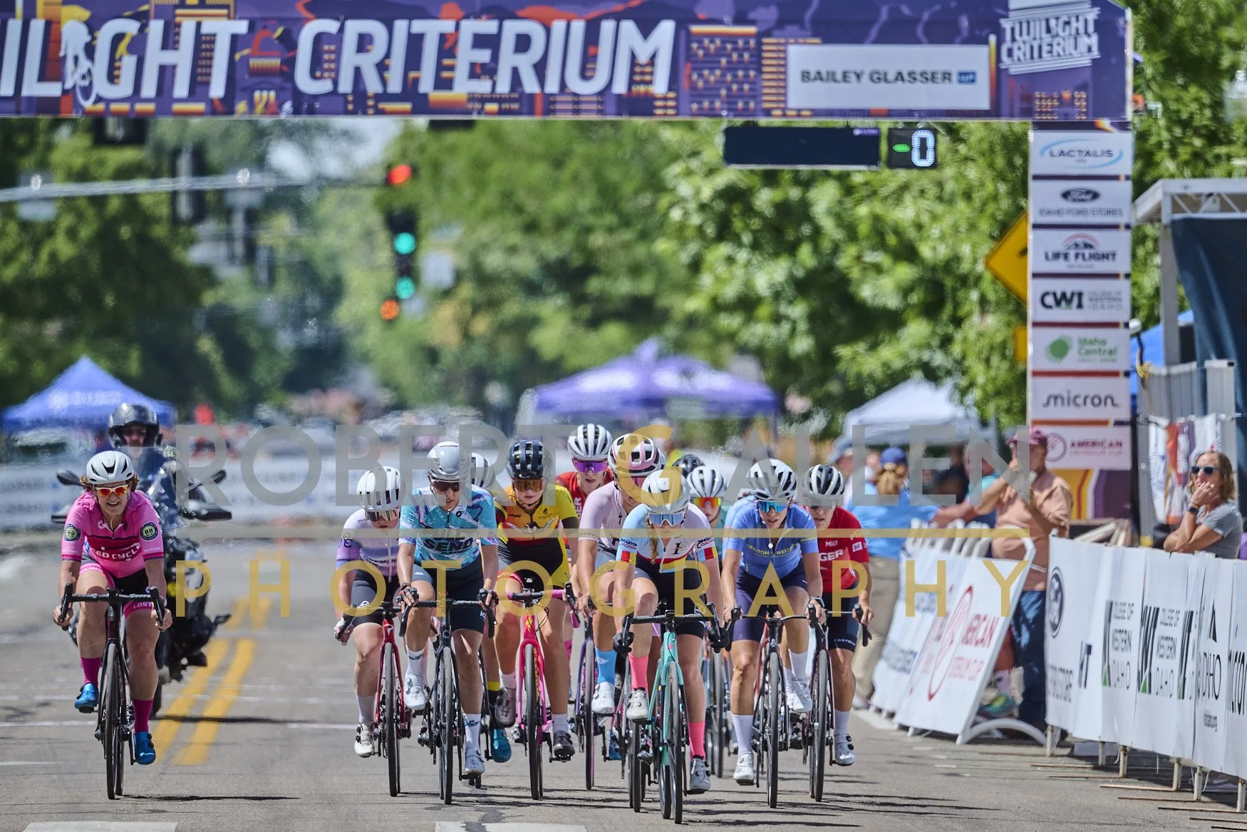 Cyclists at the start of a race, crossing the finish line under a banner reading 'Twilight Crit-era'. The event appears to be part of the Twilight Crit Series, with spectators and event tents visible on the sides.