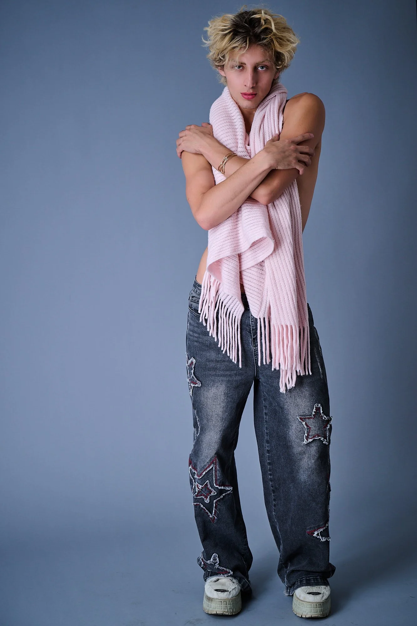 A person with short, curly blonde hair wearing a pink scarf, distressed jeans with star patches, and sneakers, standing against a blue background.