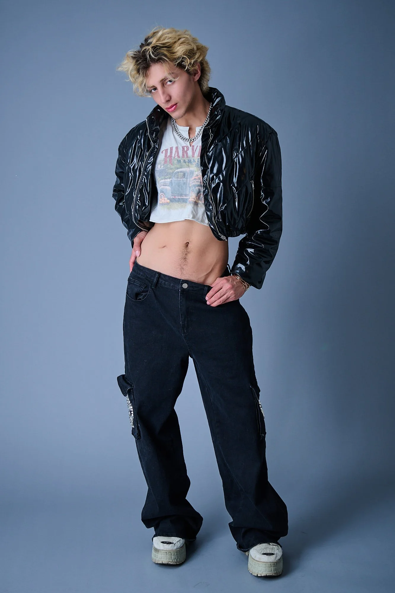 Young person with blonde curly hair, wearing a black shiny jacket, a white t-shirt with a graphic, loose black pants, and white shoes, standing against a plain gray background.