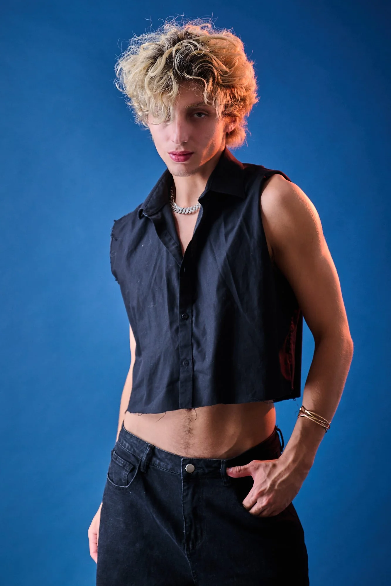 Young man with curly blonde hair wearing a sleeveless black vest, necklace, and bracelets, posing against a blue background.