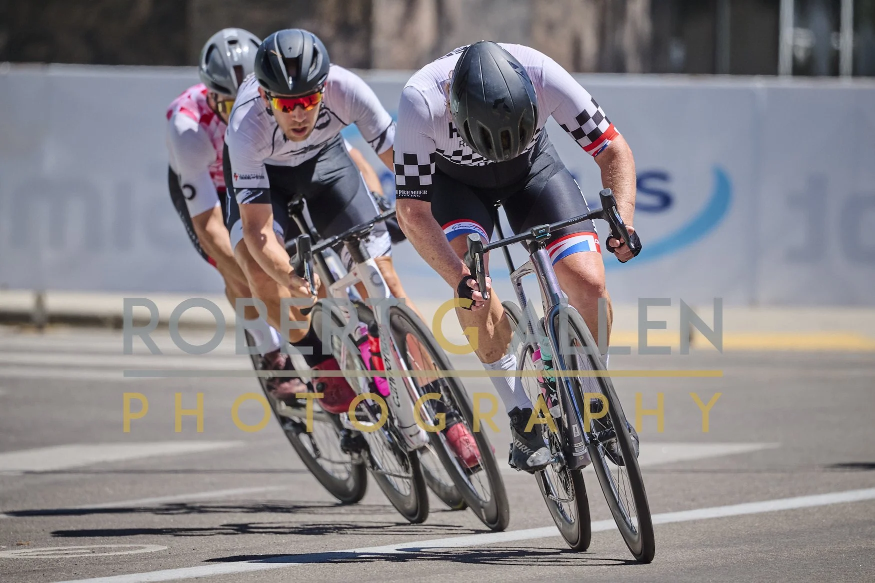 Three cyclists racing on a road, leaning into a turn, wearing helmets and cycling gear.