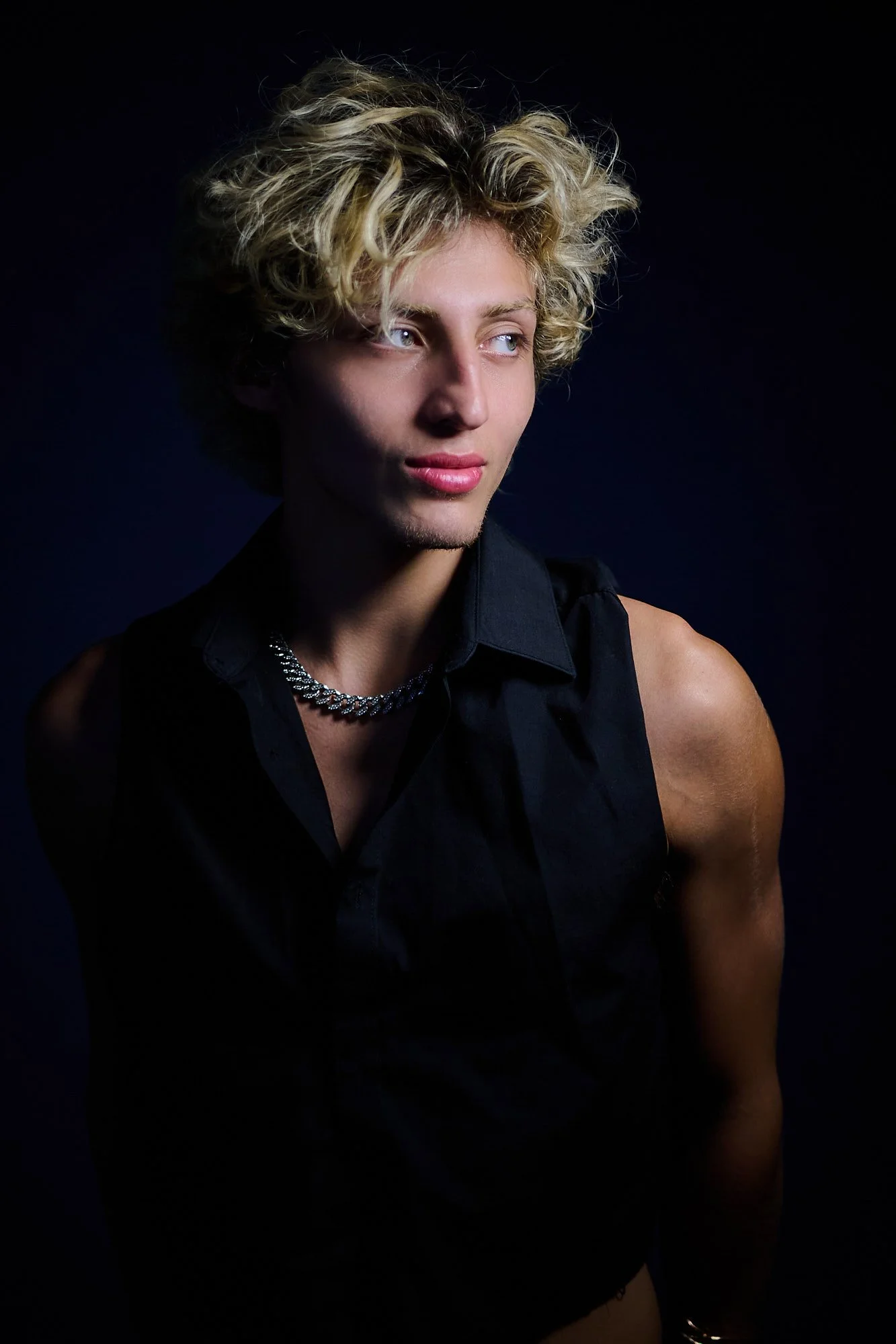 A young man with curly blonde hair and light skin wearing a sleeveless black shirt, a chunky silver chain necklace, and a silver bracelet, standing against a dark background, looking slightly to the side.