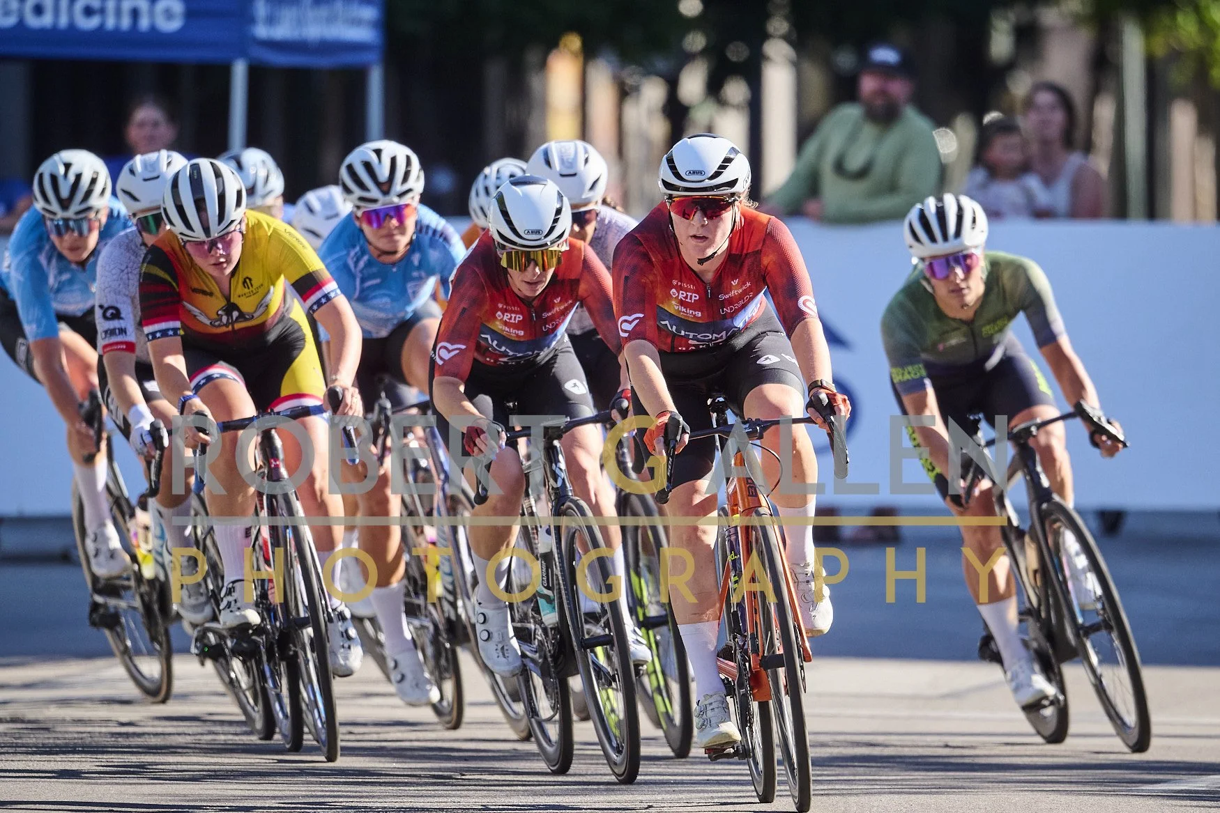 A group of competitive cyclists wearing helmets and sunglasses riding on a paved road during daytime, participating in a race or event.