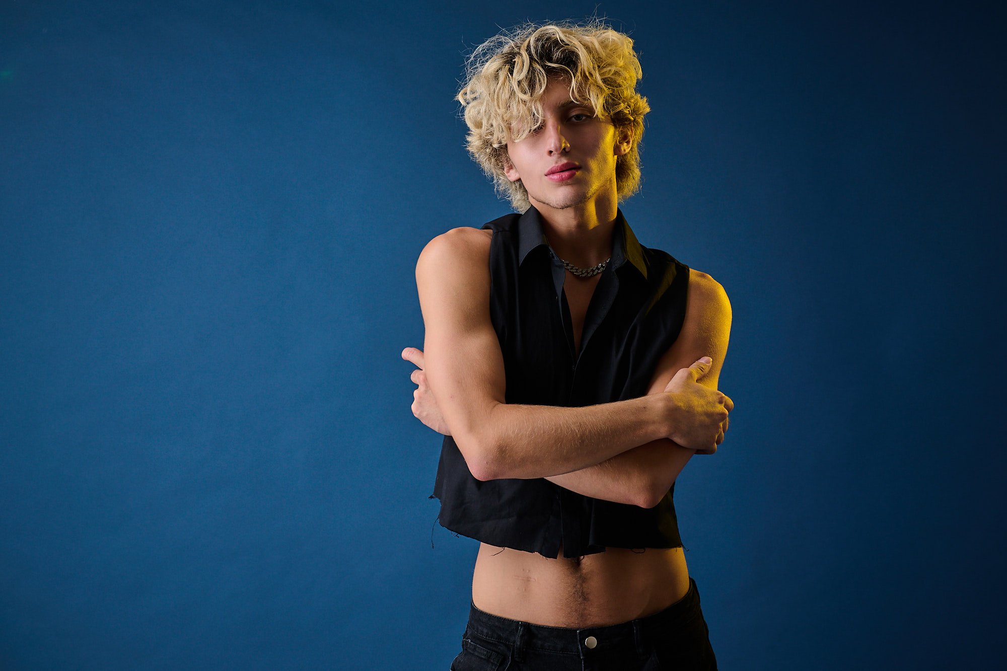 A person with curly blonde hair wearing a black sleeveless shirt and black jeans, standing against a dark blue background. They are crossing their arms and looking at the camera with a neutral expression.