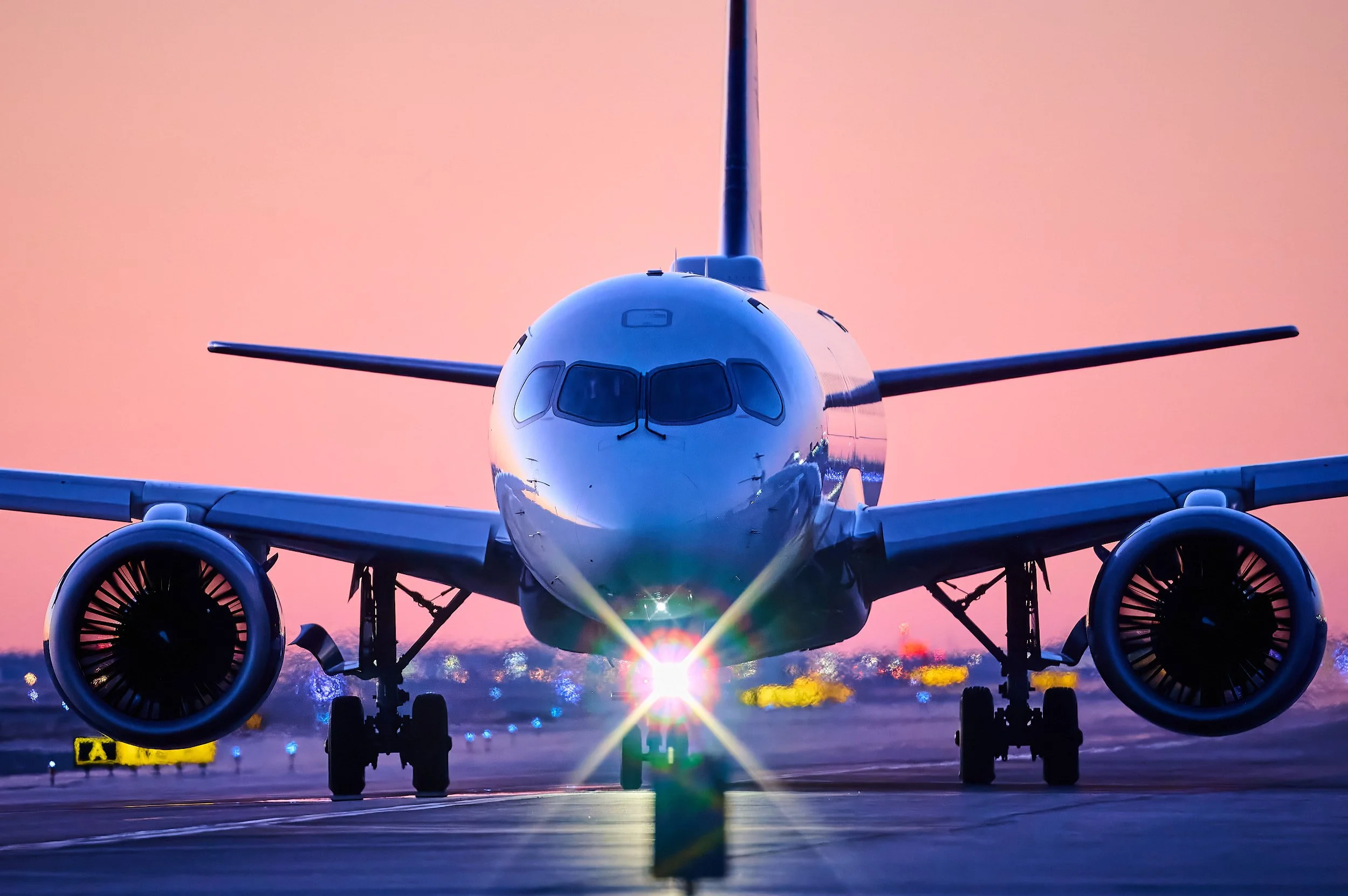 Front view of a commercial airplane on the tarmac at sunset, with a pink sky and sunlight reflecting off the aircraft's nose and wheels.