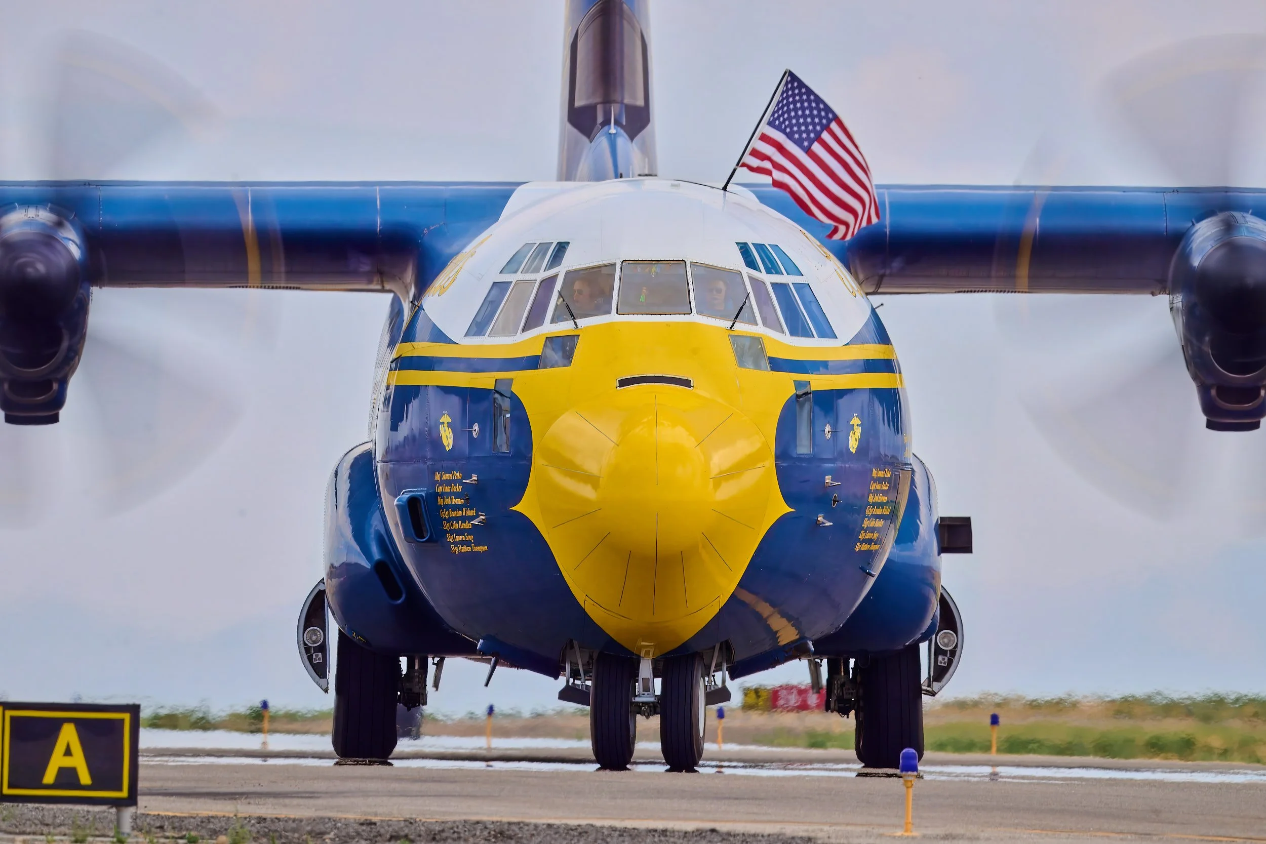 Front view of a NASA space shuttle aircraft with a yellow nose, blue wings, and an American flag on the right wingtip, situated on an airfield.