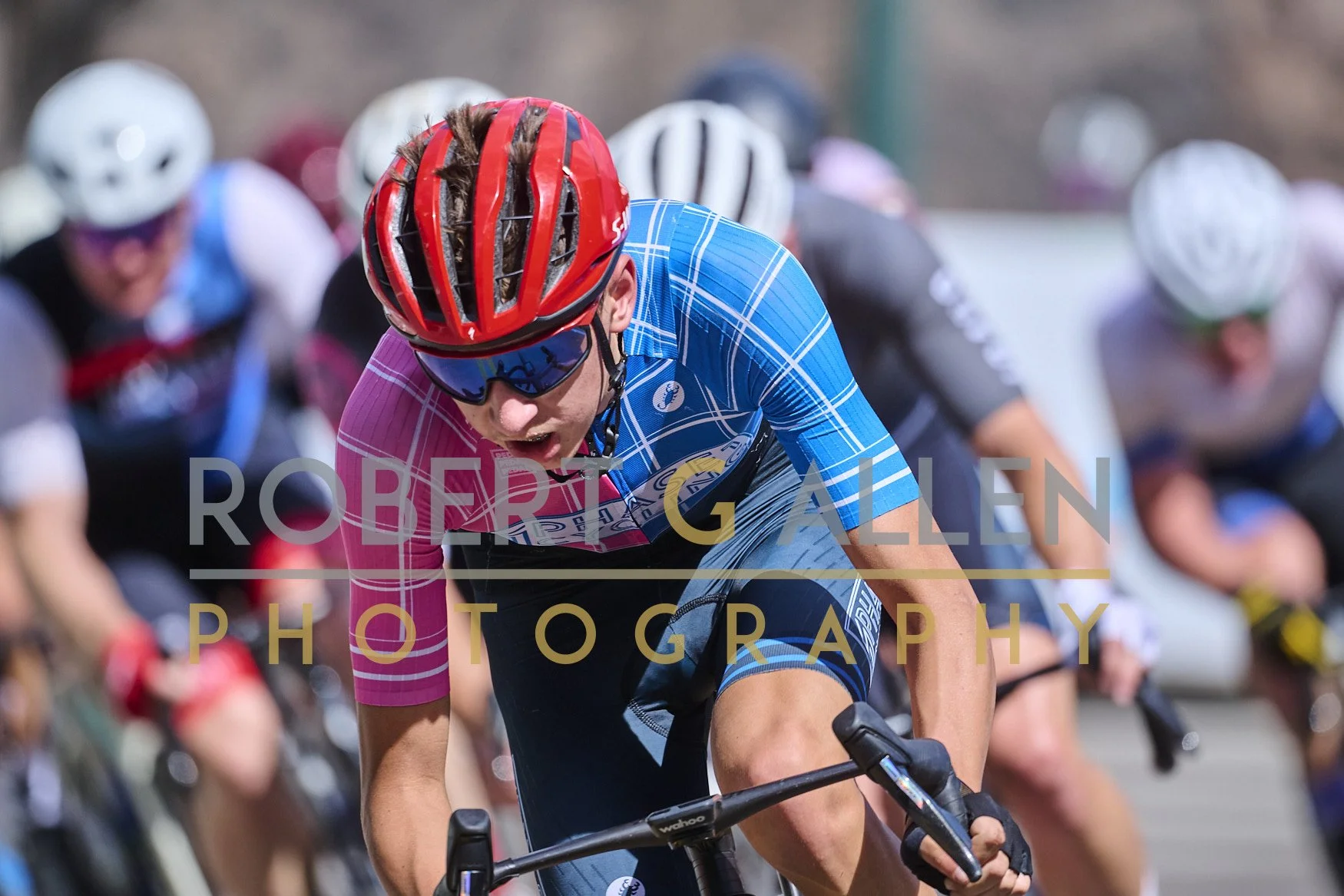 Cyclist wearing a red helmet and blue cycling outfit participating in a race with other cyclists in the background.