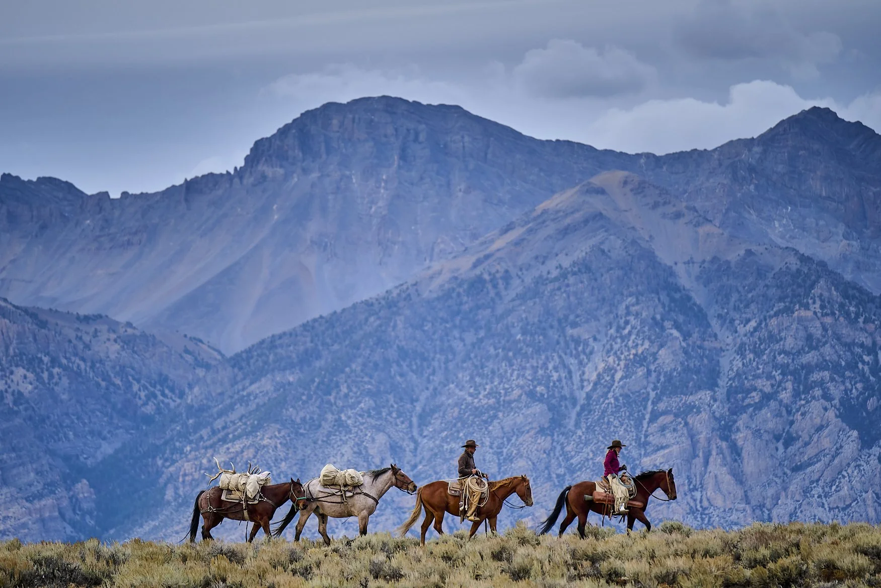 A group of people riding horses through a landscape with mountains in the background, under a cloudy sky.