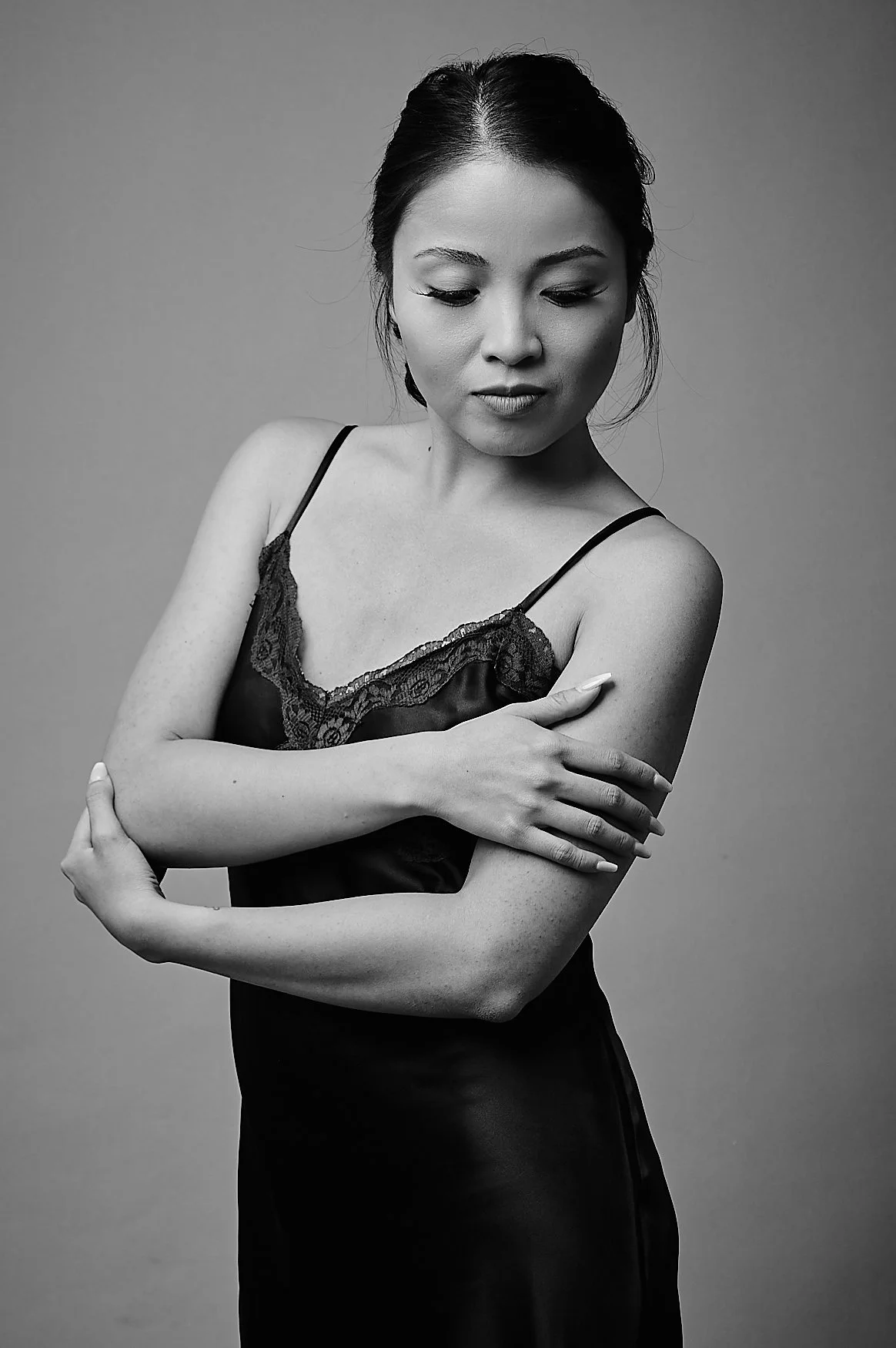 A woman in a satin and lace slip dress looks down with crossed arms, in a black-and-white studio portrait.