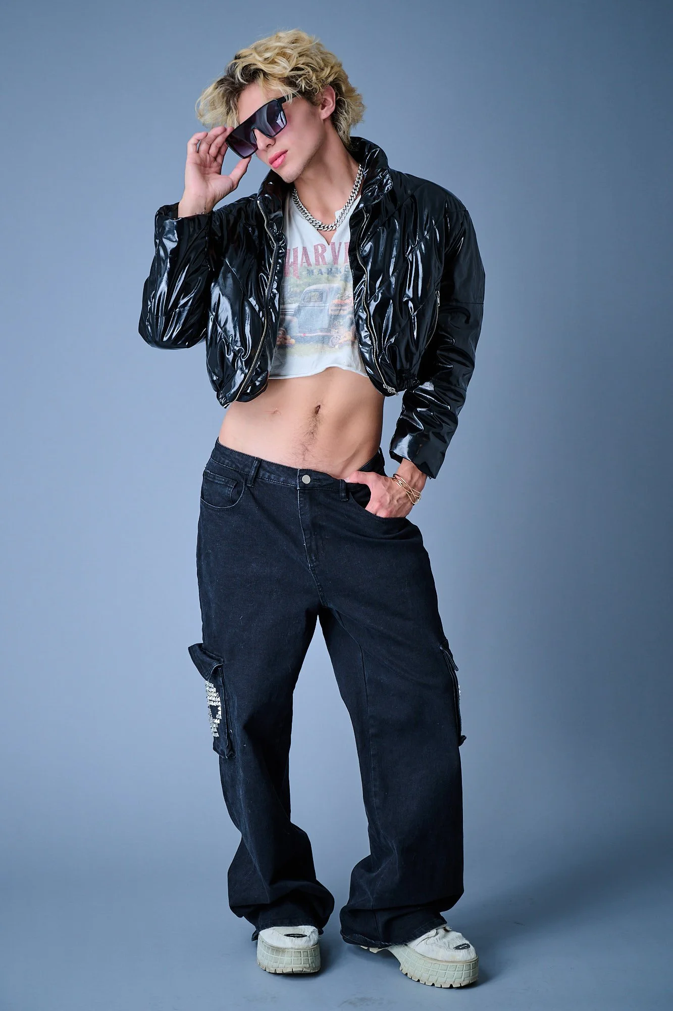 Person with blonde, curly hair wearing dark sunglasses, a black shiny jacket, a vintage graphic T-shirt, baggy black cargo pants, and chunky sneakers, posing against a plain gray background.