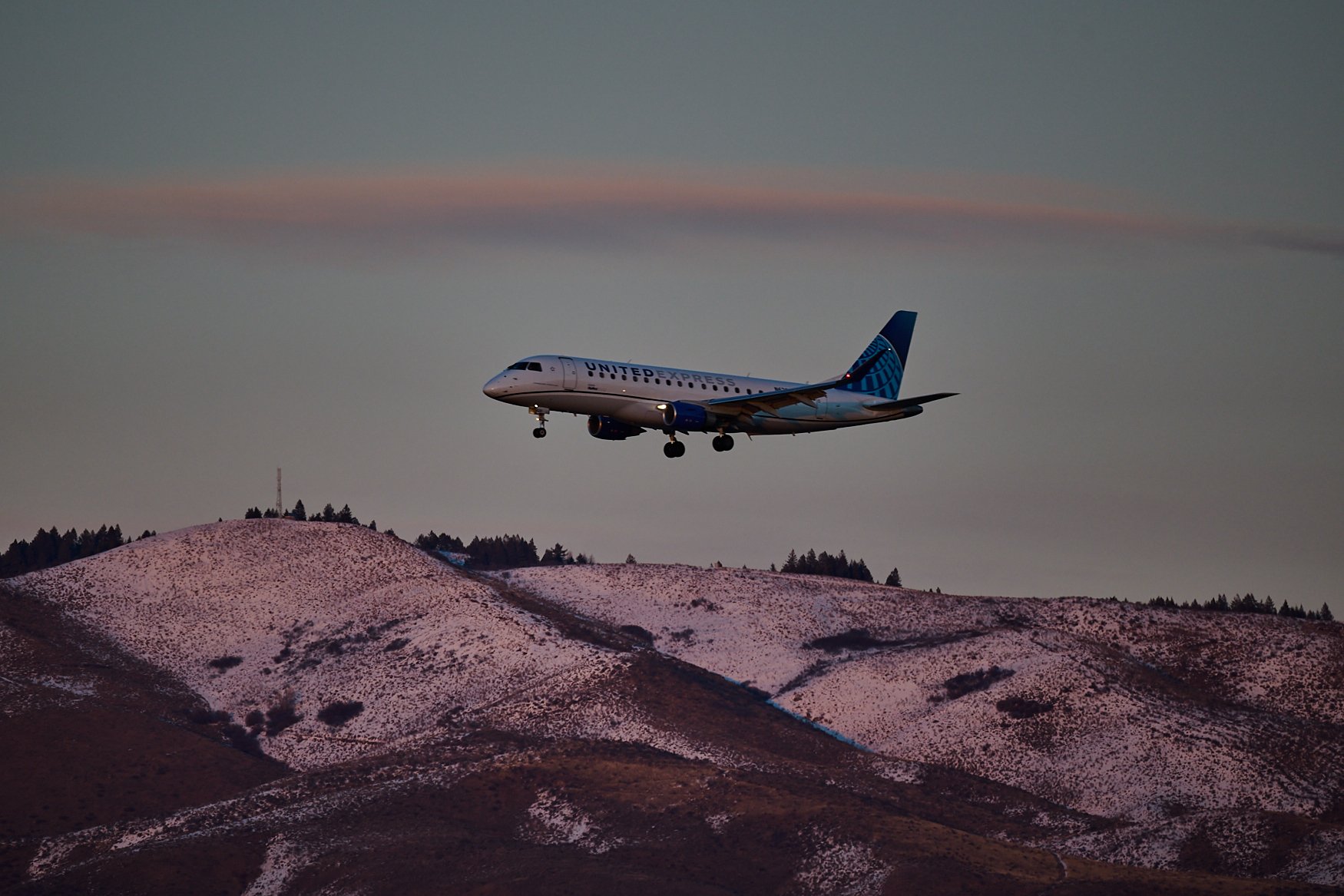 An airplane with United Express branding flying over snowy hills at sunset.