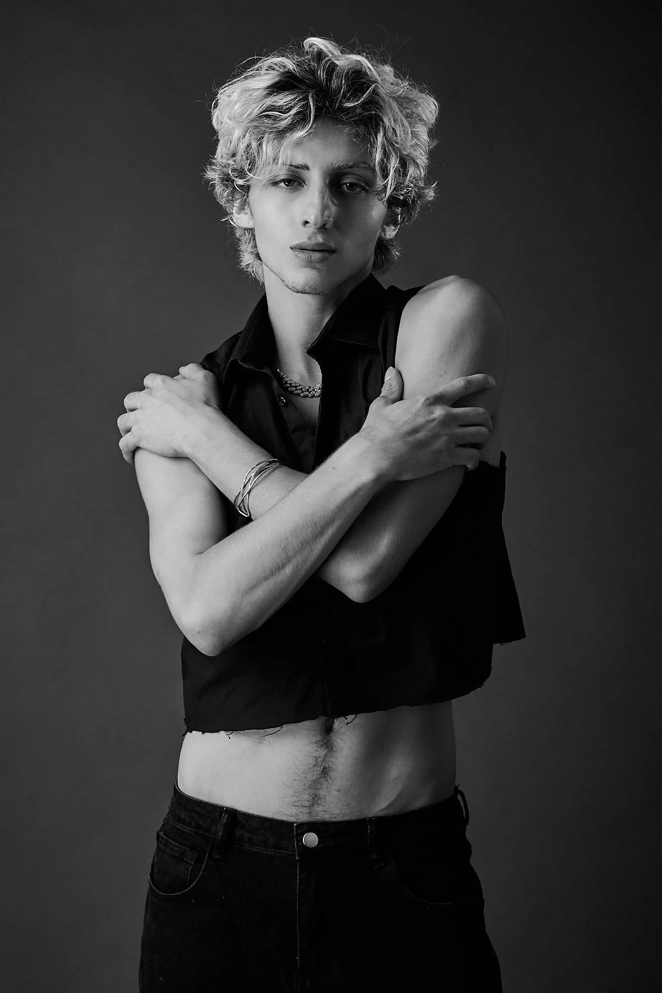 Black and white photo of a person with short, curly blonde hair, wearing a sleeveless black shirt and black jeans, standing against a plain dark background with arms crossed over their chest.