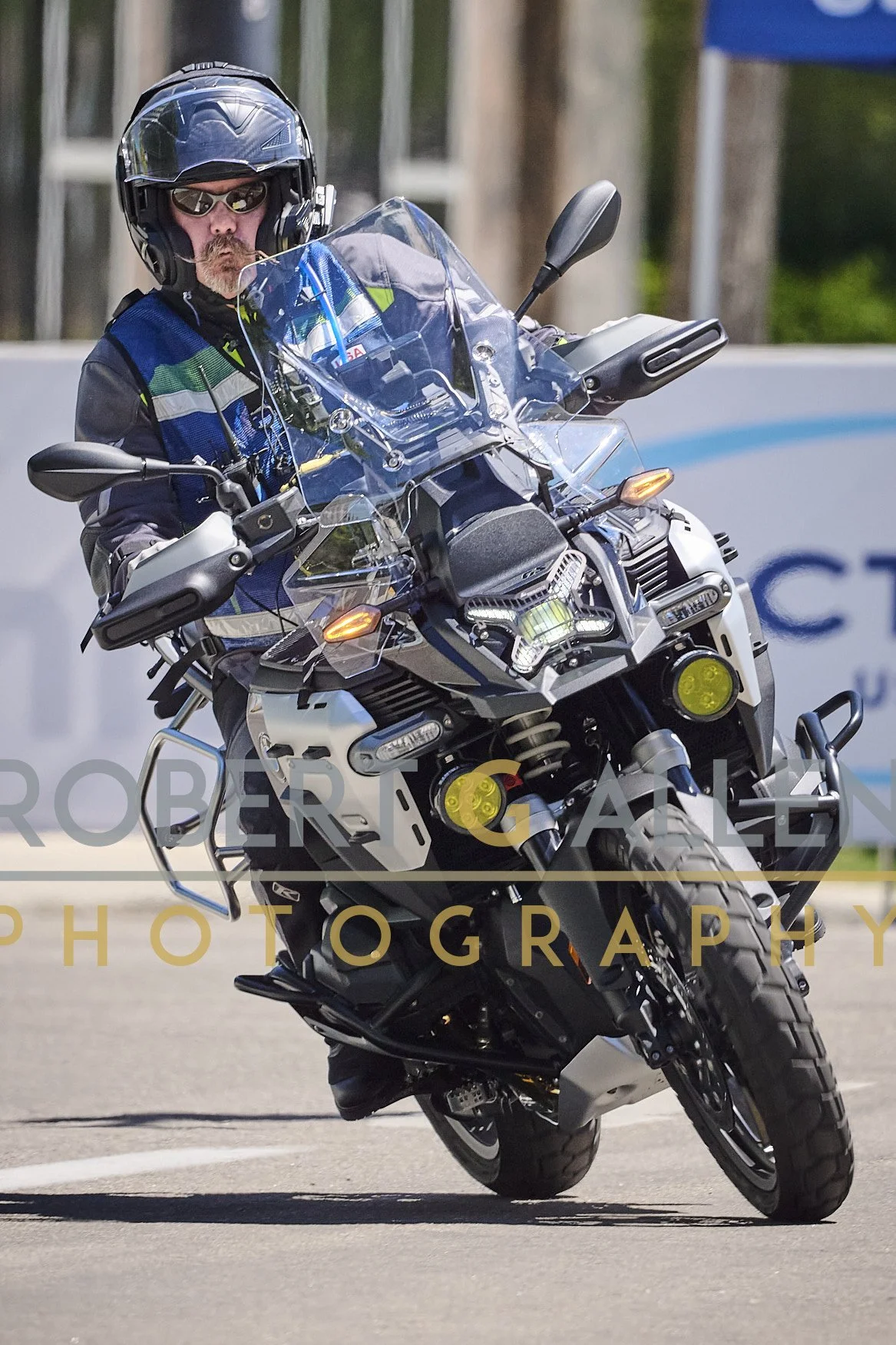 A person riding an adventure motorcycle, wearing a helmet and sunglasses, performing a wheelie on a paved road with trees in the background.