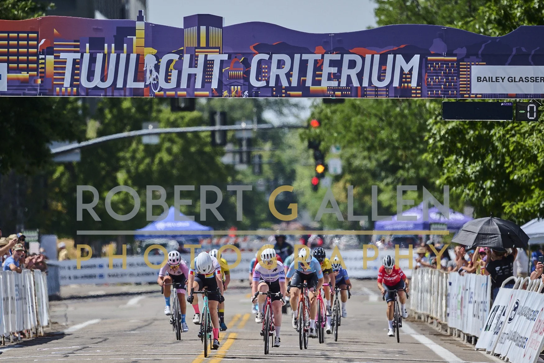 Cyclists competing in a race crossing the finish line at the Twilight Critérium with spectators on both sides and a cityscape banner overhead.