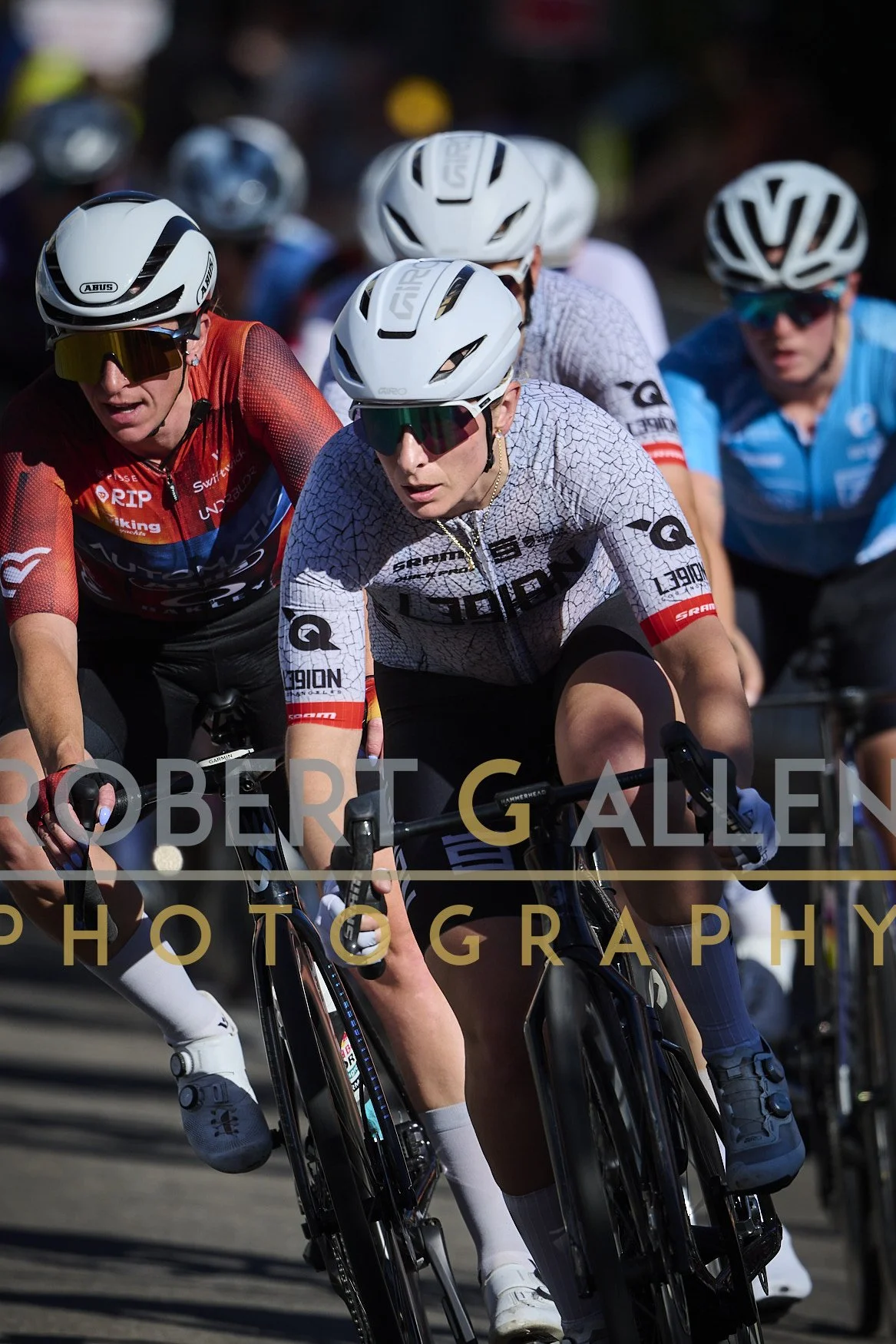 Group of female cyclists racing, wearing helmets and sports sunglasses, in an outdoor setting.