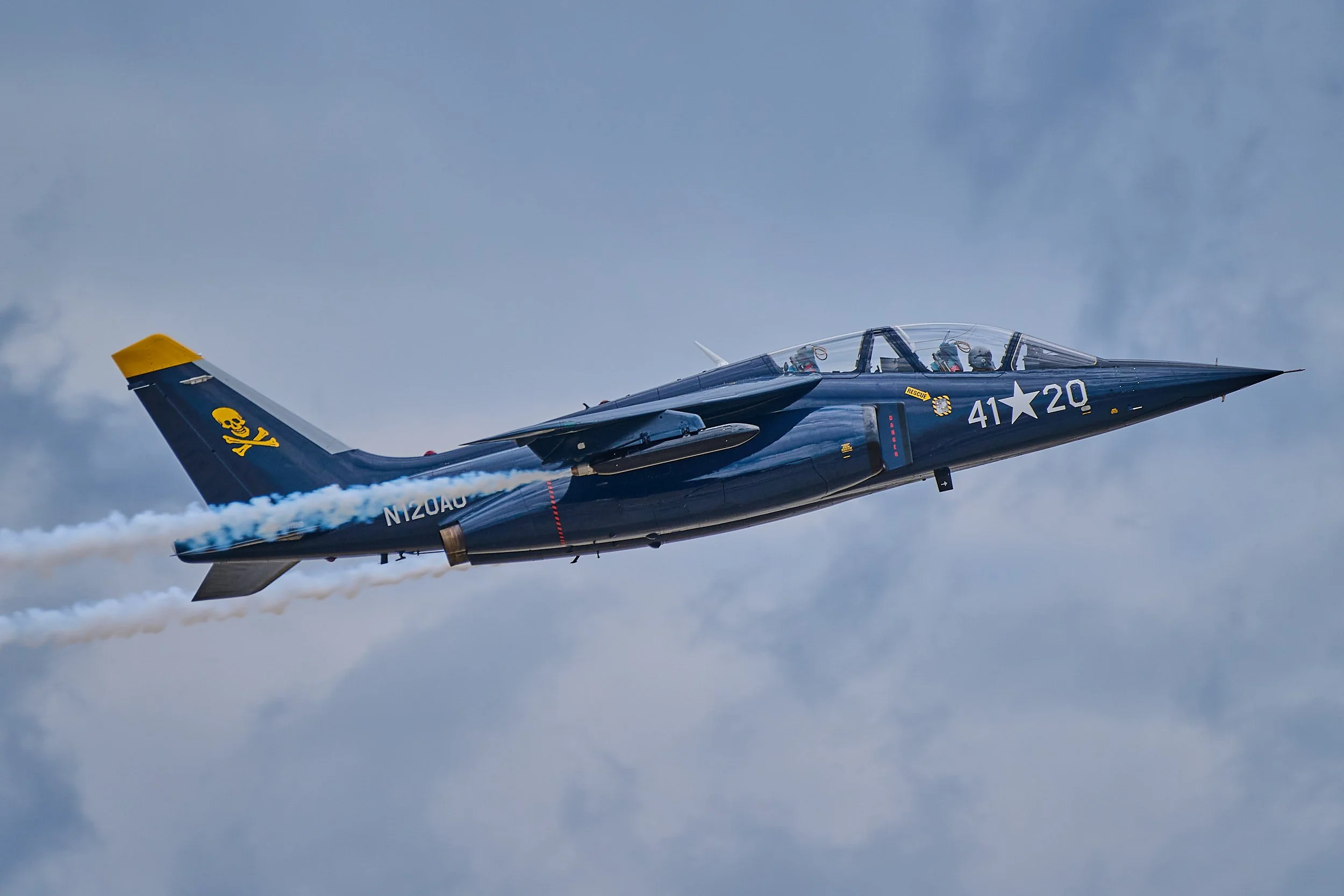 A jet aircraft with blue and gray camouflage, yellow tail fin with a skull and crossbones, flying through the sky, emitting white smoke.