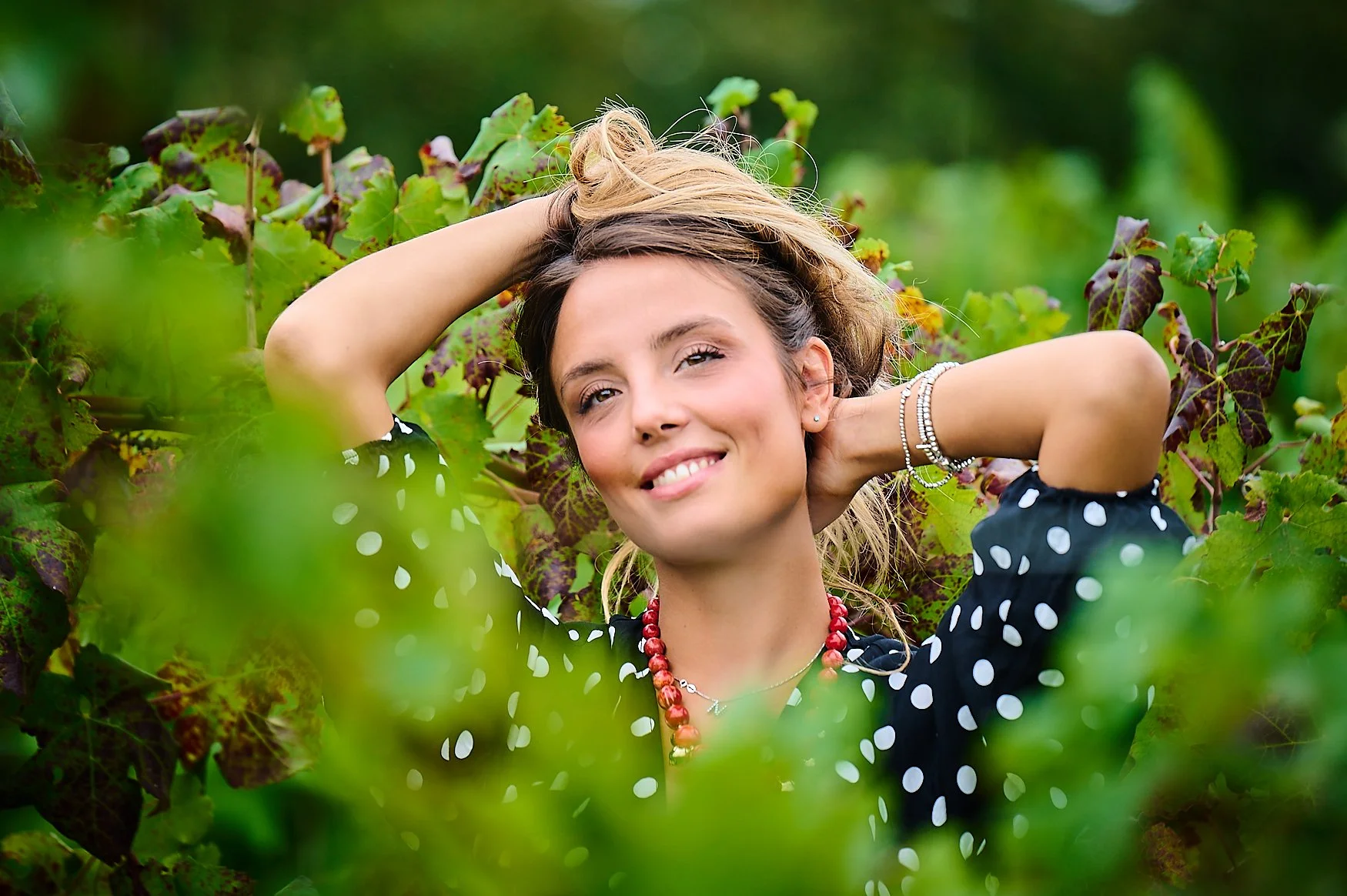A woman with blonde hair and light skin, smiling, with one hand behind her head, wearing a black polka dot dress and jewelry, standing among green and purple foliage.