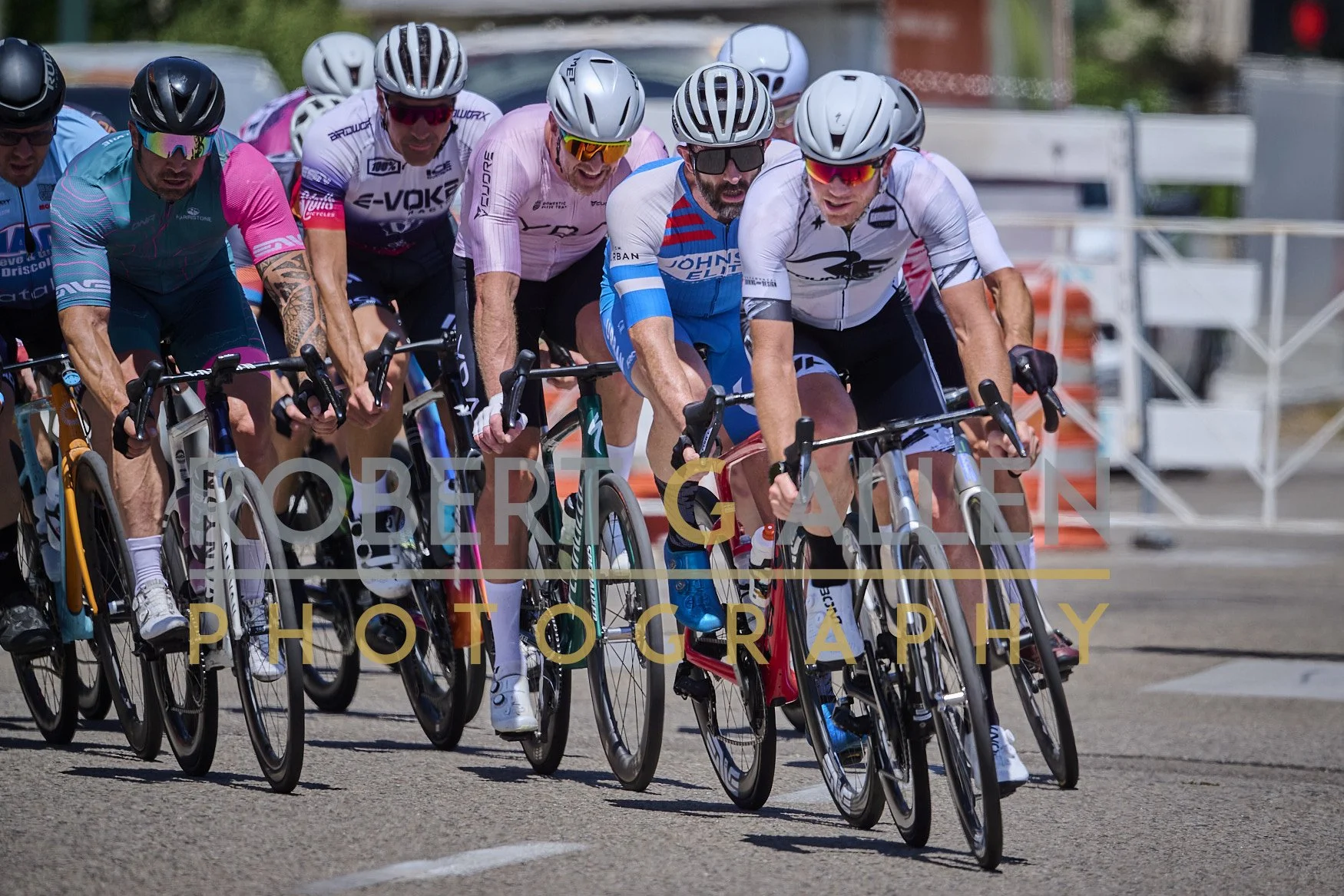 A group of cyclists racing on a road, wearing helmets and colorful jerseys, with some in sunglasses, pushing hard in a competitive bike race.