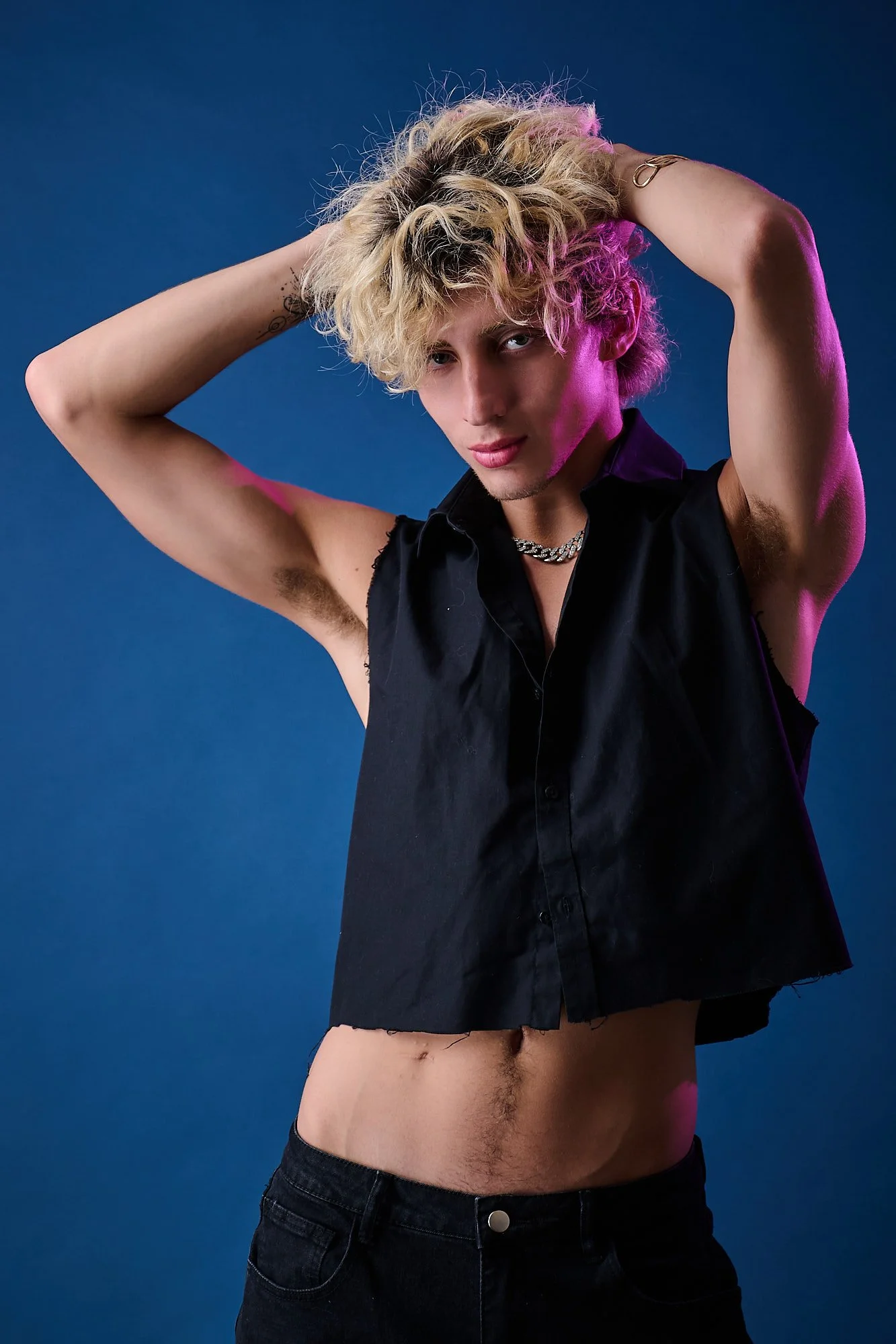 A person with short curly blonde hair and blue eyes posing with hands behind head, wearing a sleeveless black shirt, chain necklace, and black jeans, against a solid blue background, with colored lighting pink and purple highlights on face and hair.