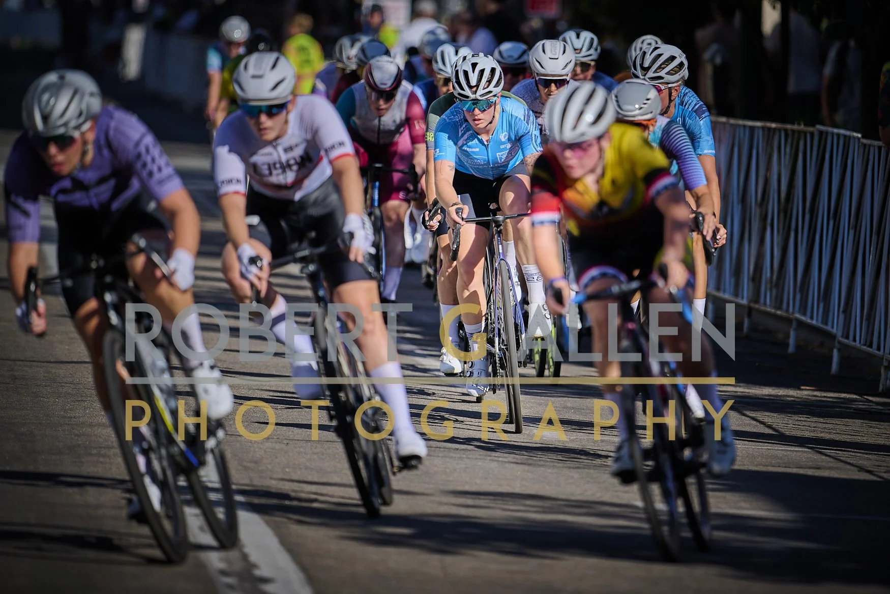 Cyclists in a race, wearing helmets and racing gear, riding on a street with barriers on the side.