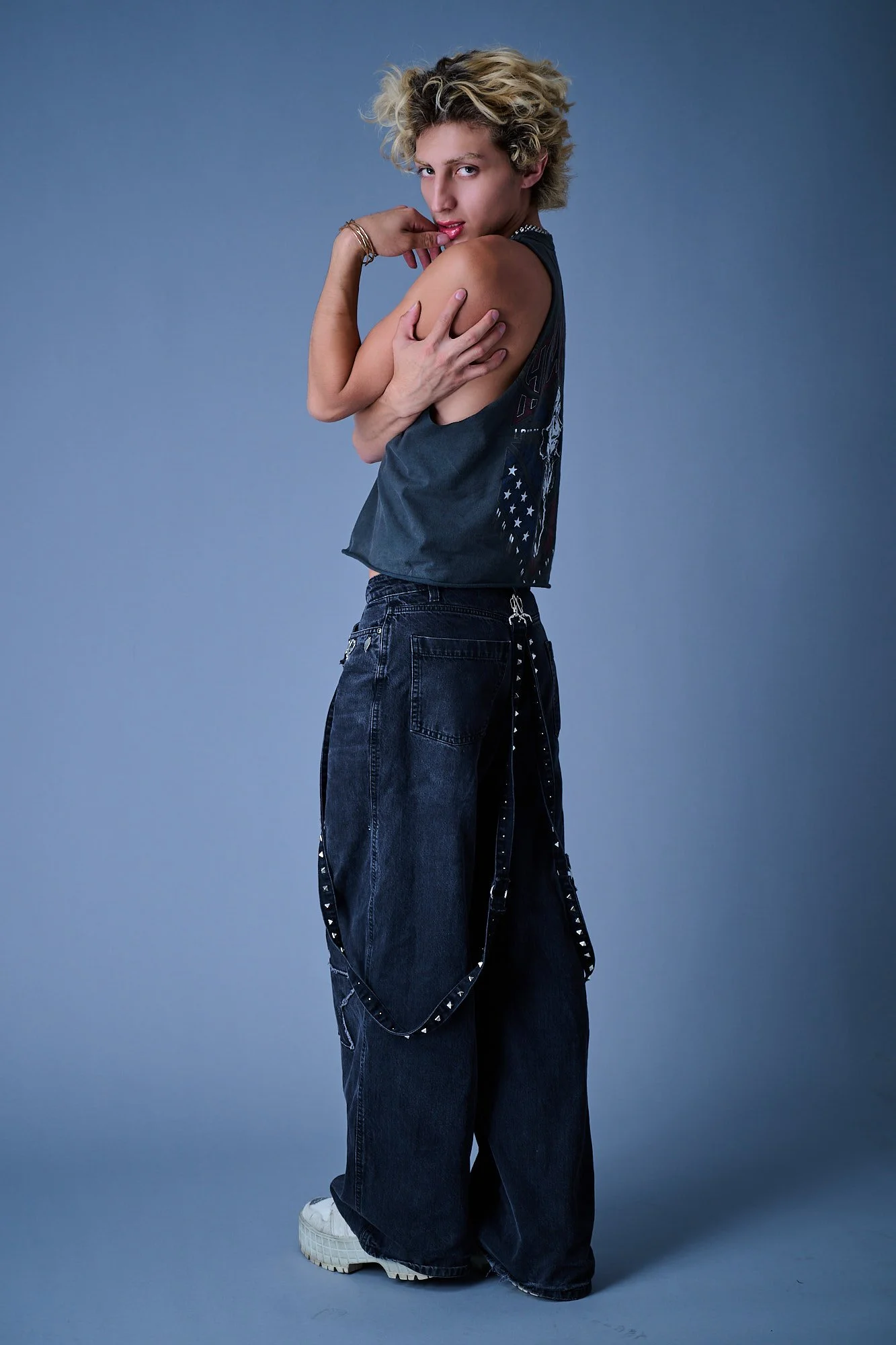 A person with blonde curly hair posing against a plain blue background, wearing a sleeveless shirt, baggy jeans with studded straps, and white sneakers.