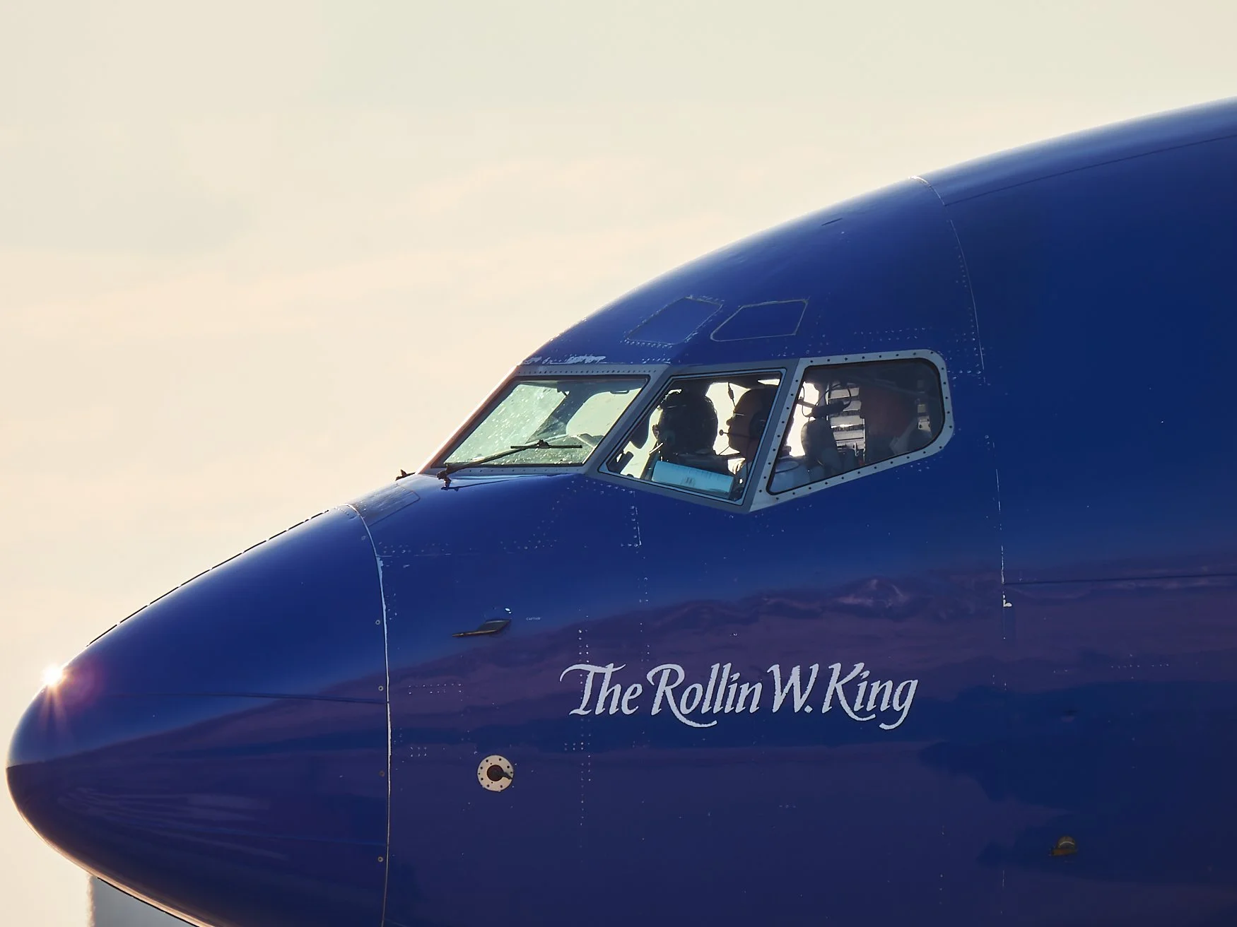 Close-up of the cockpit of a blue airplane with visible pilots, with the words "The Rollin W. King" inscribed on the side.