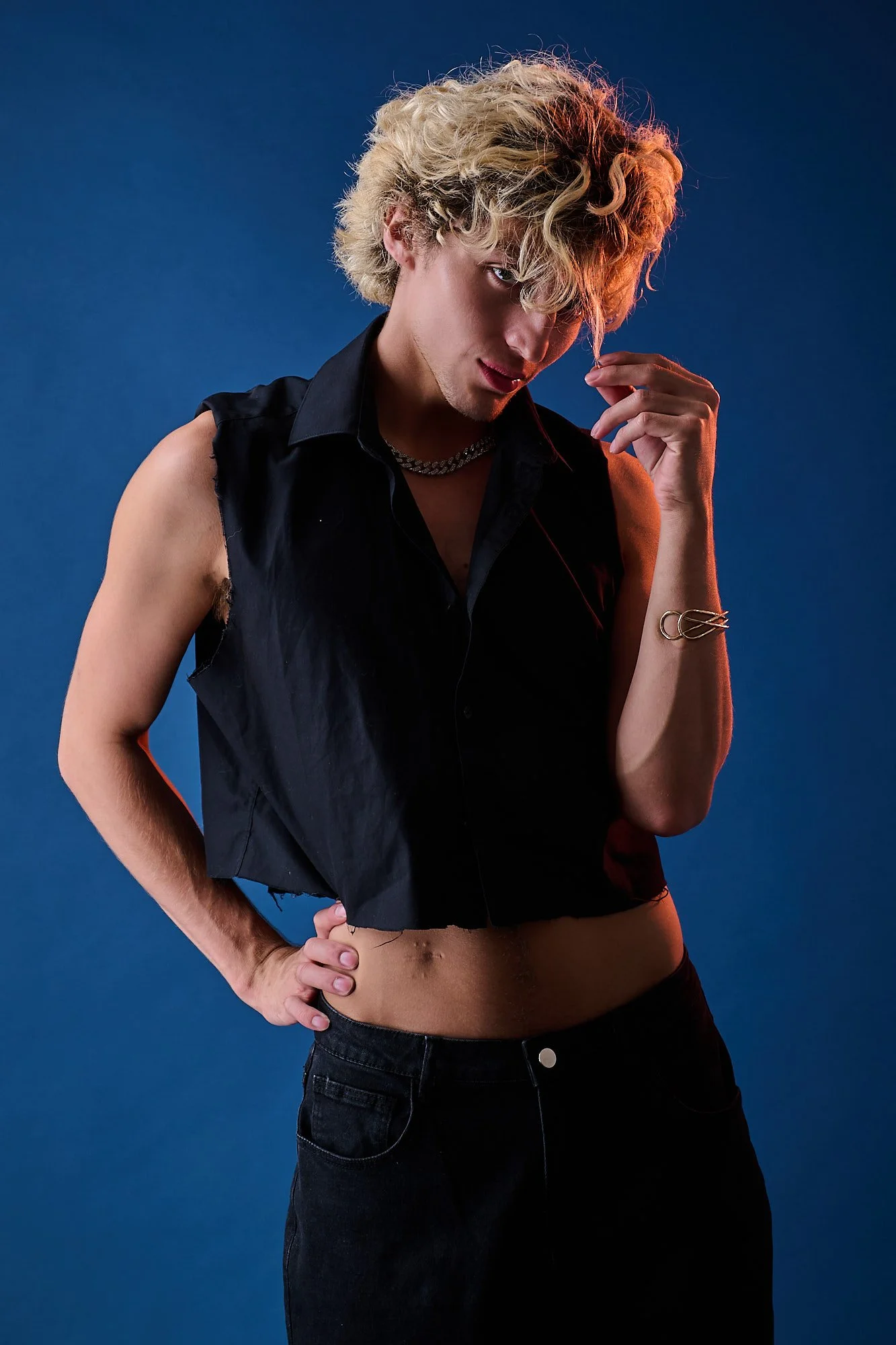 A young person with curly blonde hair, wearing a sleeveless black shirt, chain necklace, and jewelry, posing against a blue background.