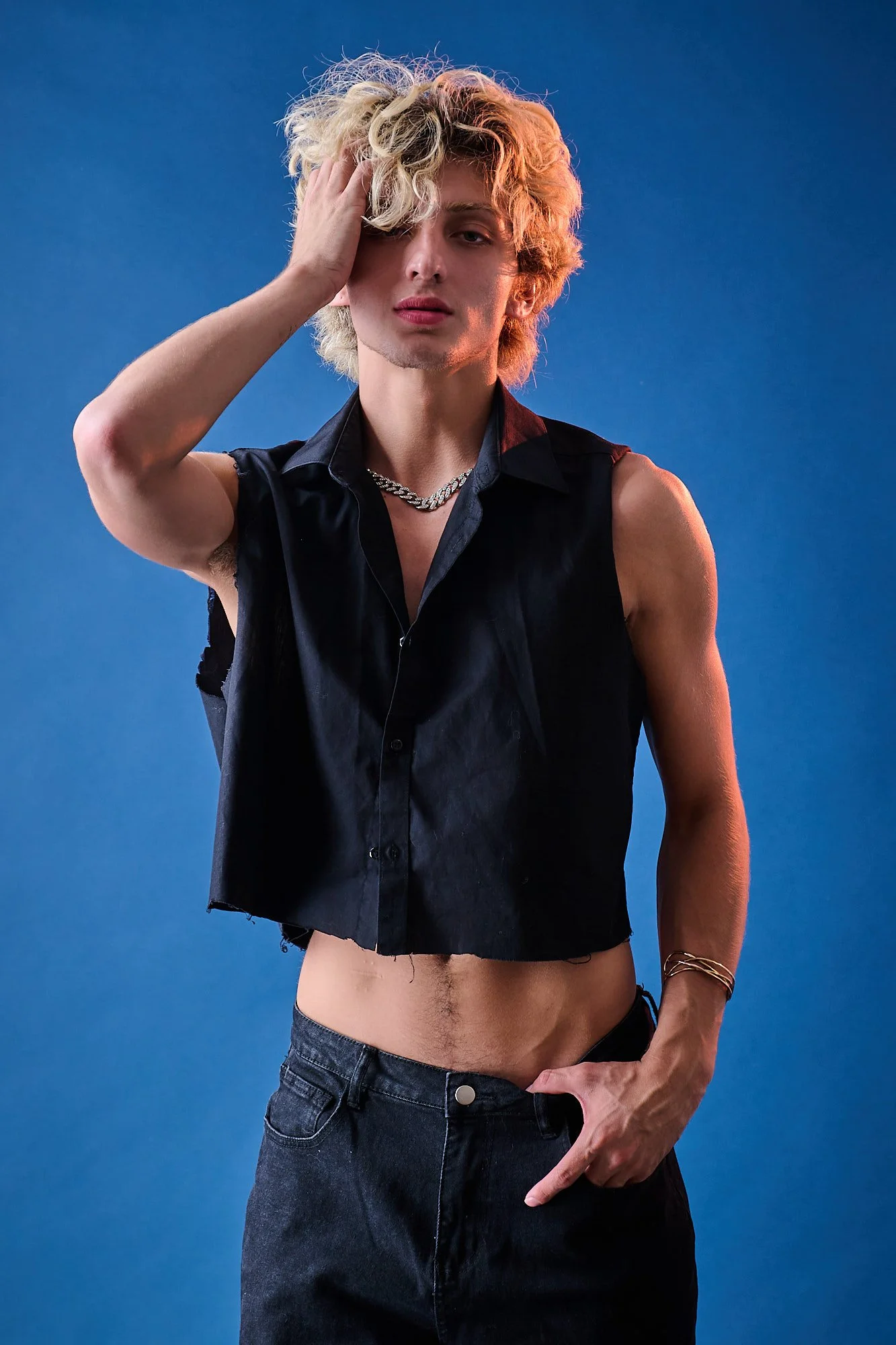 A young man with curly blonde hair and fair skin poses shirtless wearing dark jeans against a blue background. He is touching his hair with his left hand, wearing a sleeveless black vest, a thick chain necklace, and a bracelet on his right wrist.
