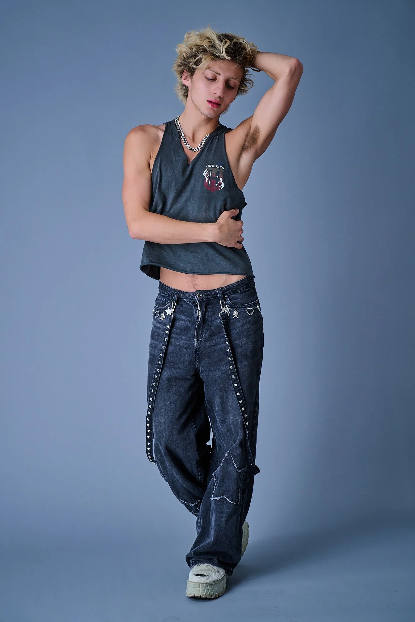 Young man with blonde curly hair posing with one arm behind his head and the other resting on his stomach. He's wearing a sleeveless black shirt with a logo, baggy jeans with decorative stripes, and white sneakers, against a plain blue background.