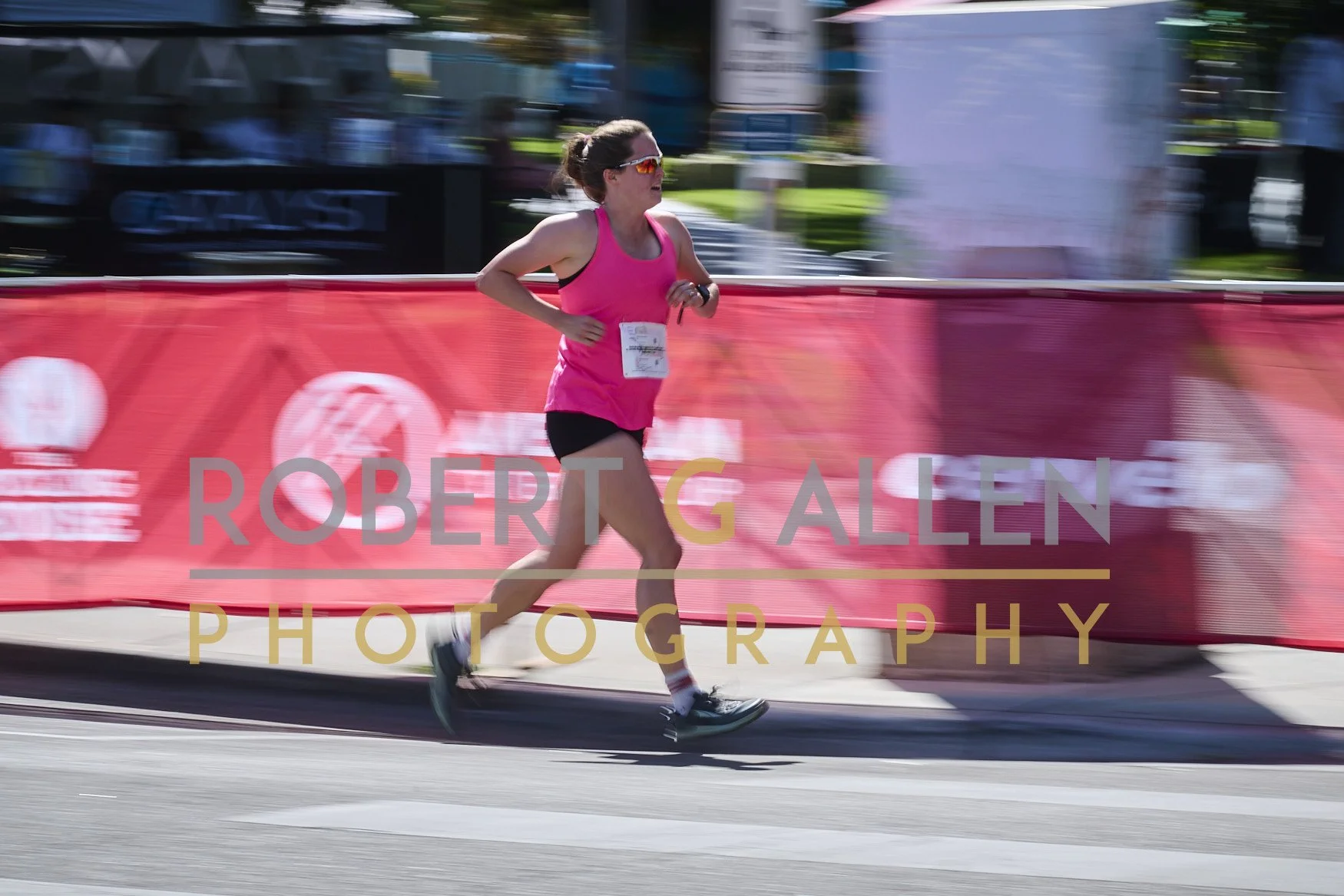 A woman running in a marathon wearing pink shirt, black shorts, sunglasses, and a race bib, passing red barriers with the text 'Robert G Allen Photography' during daytime.