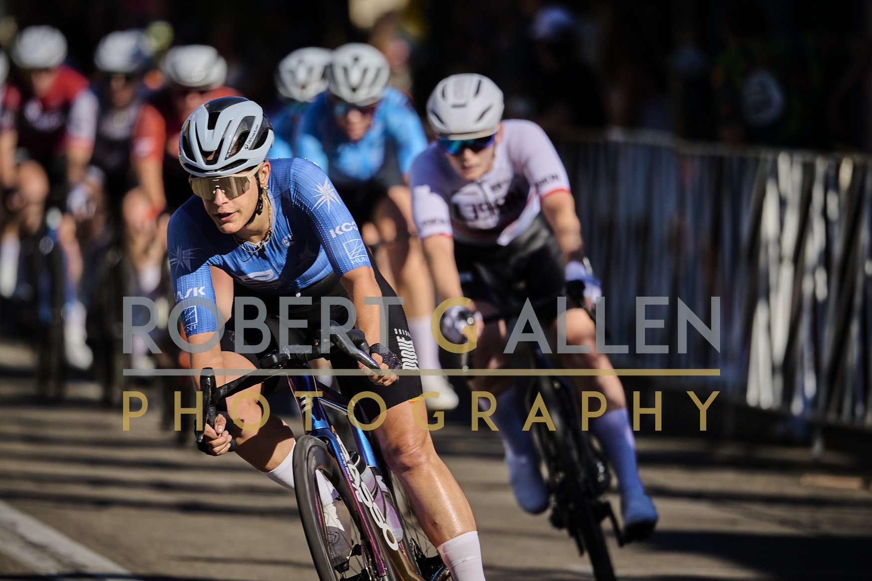 Cyclists racing on a street, with the focus on a female cyclist in a blue kit and helmet, leading the pack.