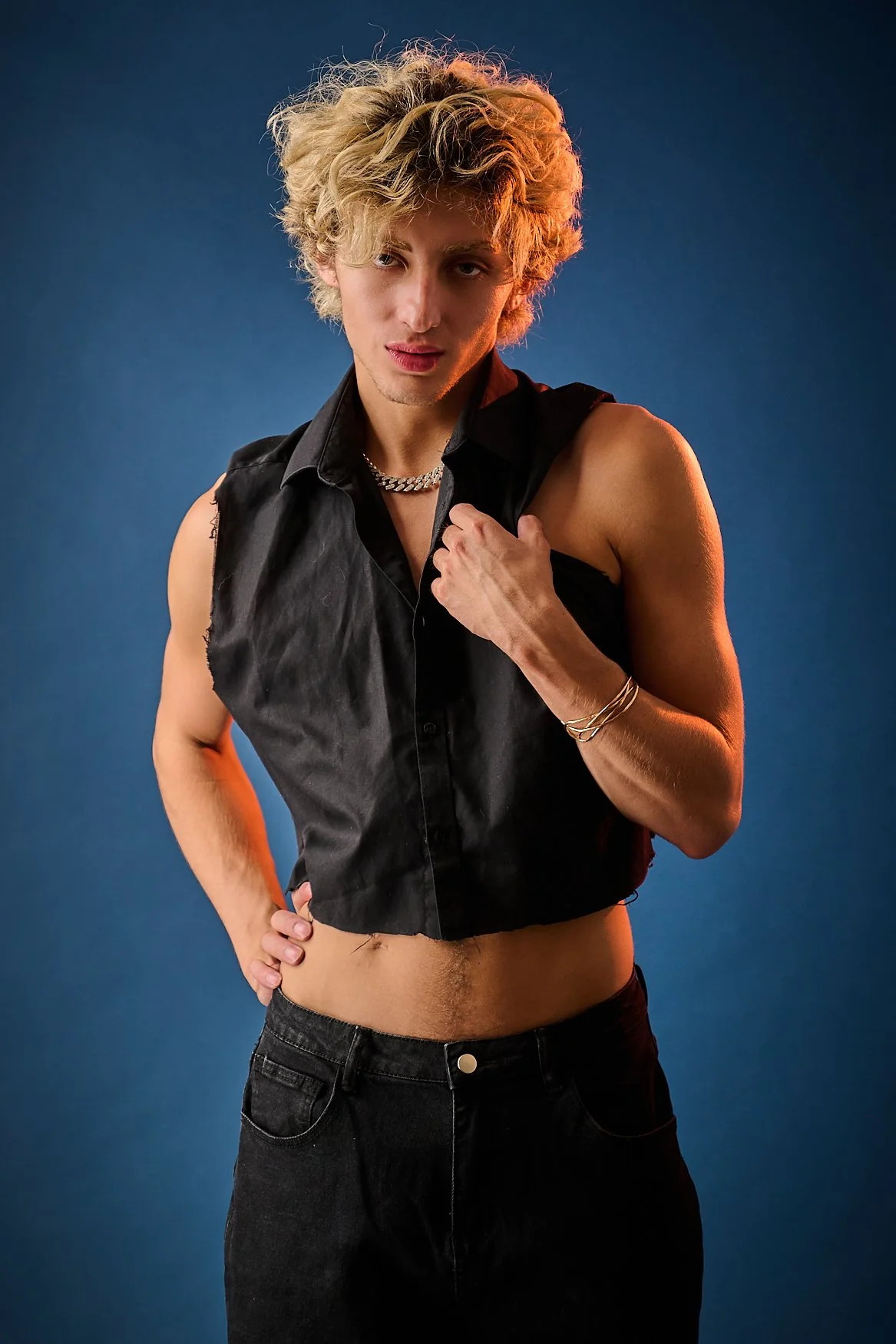 Young man with curly blond hair, wearing a sleeveless black shirt, gold chain, and bracelets, standing against a blue background, with one hand on his hip and the other touching his shirt.