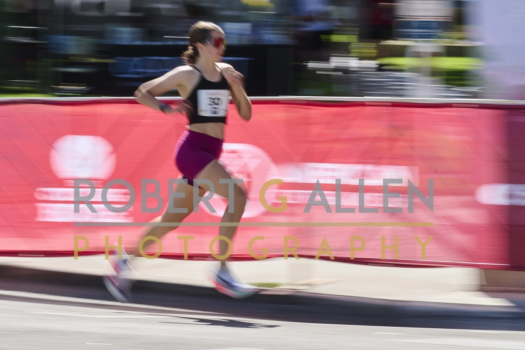 A female runner wearing sunglasses and a sports outfit runs in a race, with a red barrier displaying the text 'ROBERT GALLEN PHOTOGRAPHY' in the foreground.