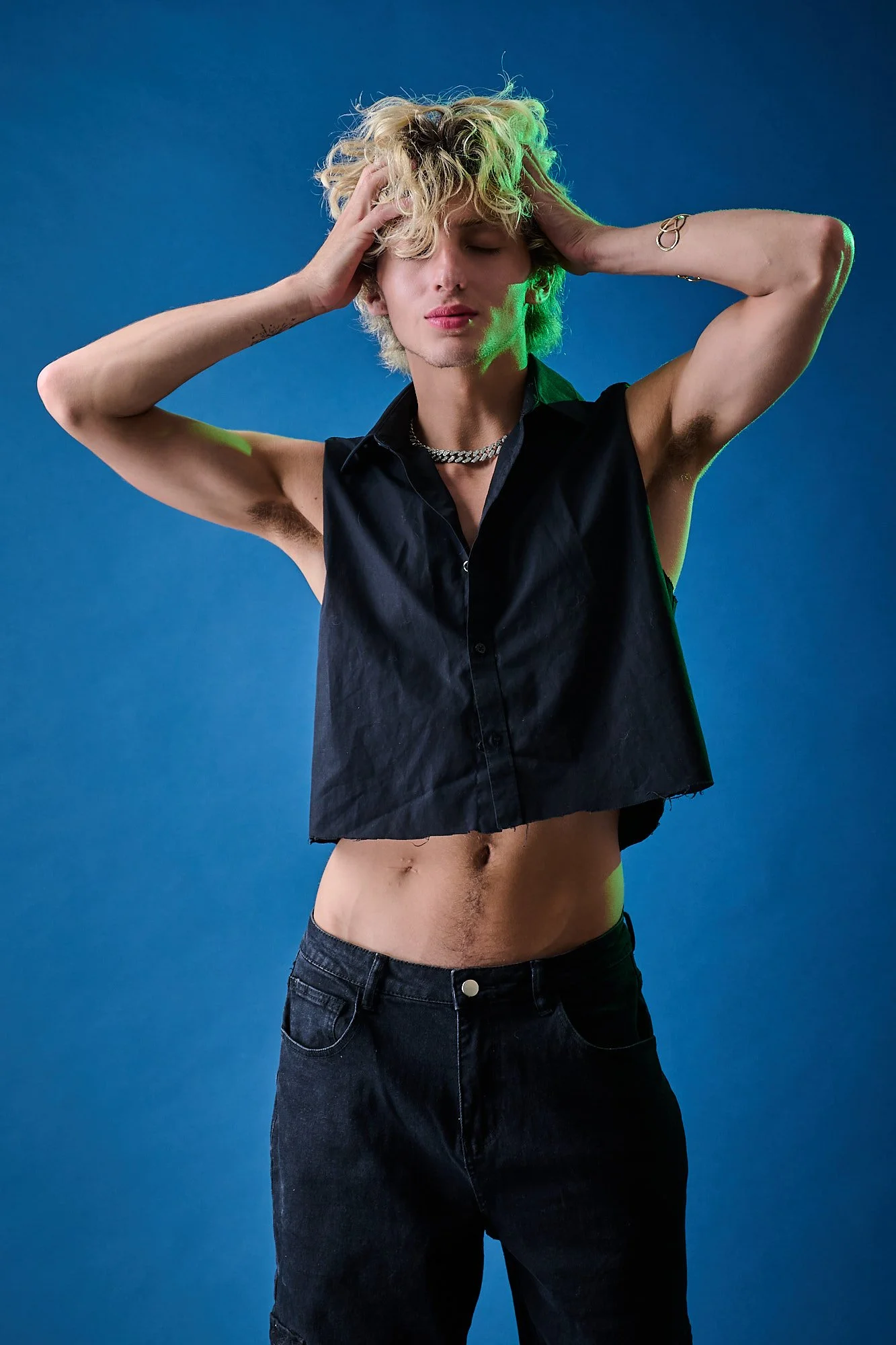 Young man with blonde curly hair poses against a blue background, wearing a sleeveless black shirt and black jeans with no shirt underneath, with his eyes closed and hands on his head.
